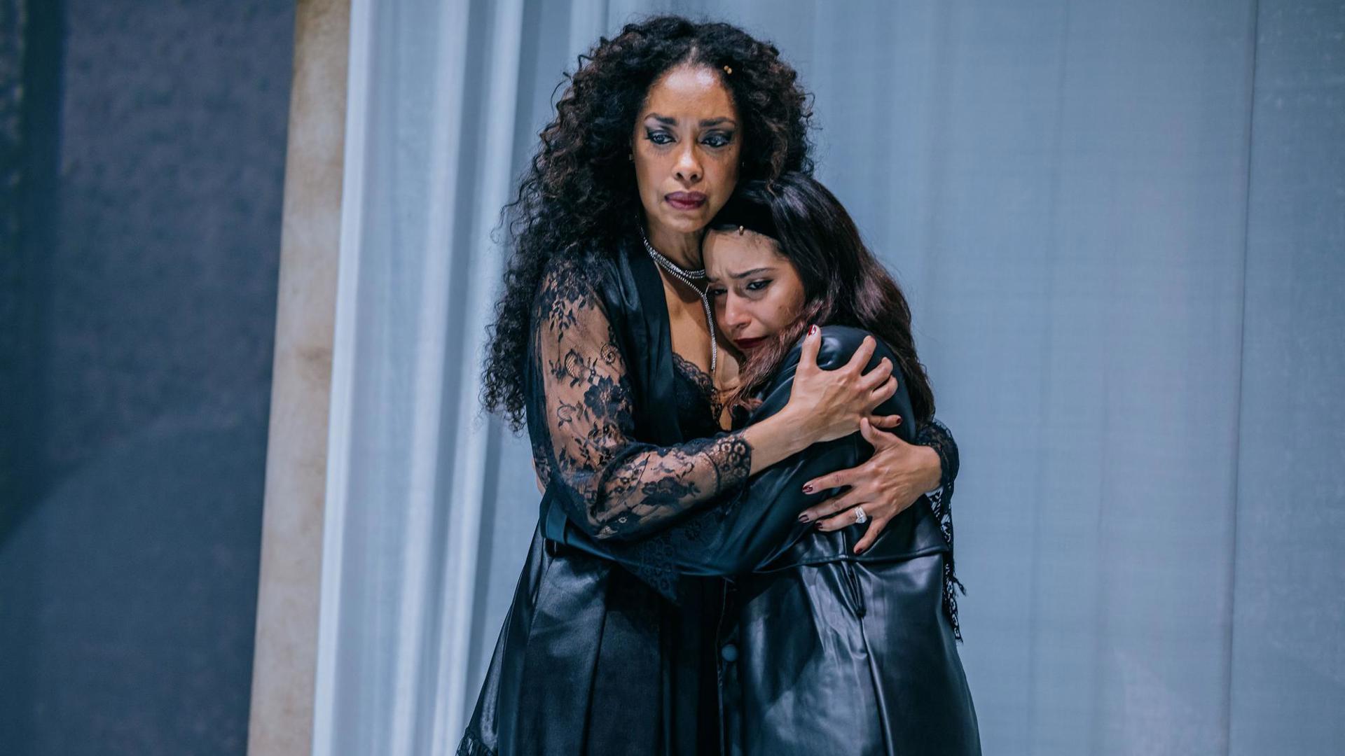Two actors embrace with solemn expressions.