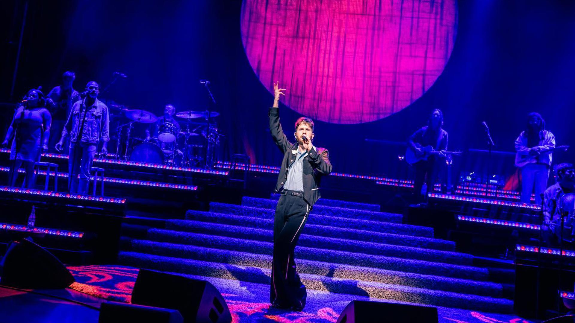 Ben Platt on stage with band and backup vocalists under blue and pink lighting with a large circular backdrop.