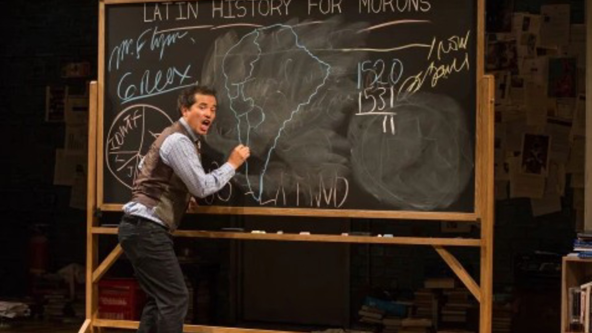 John Leguizamo writing on a chalkboard for Latin History For Morons