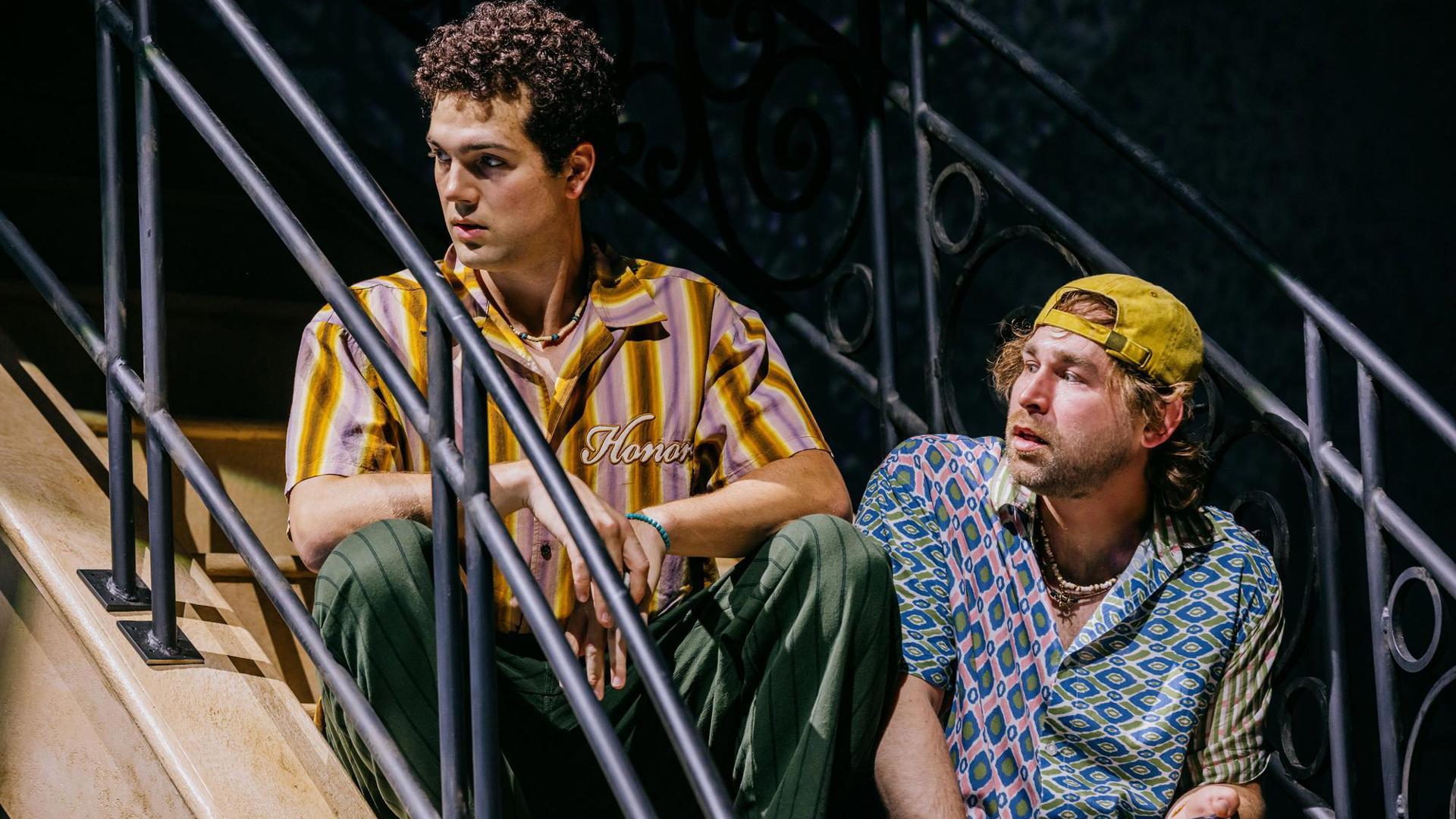 Two actors sit on a staircase, looking to the left.