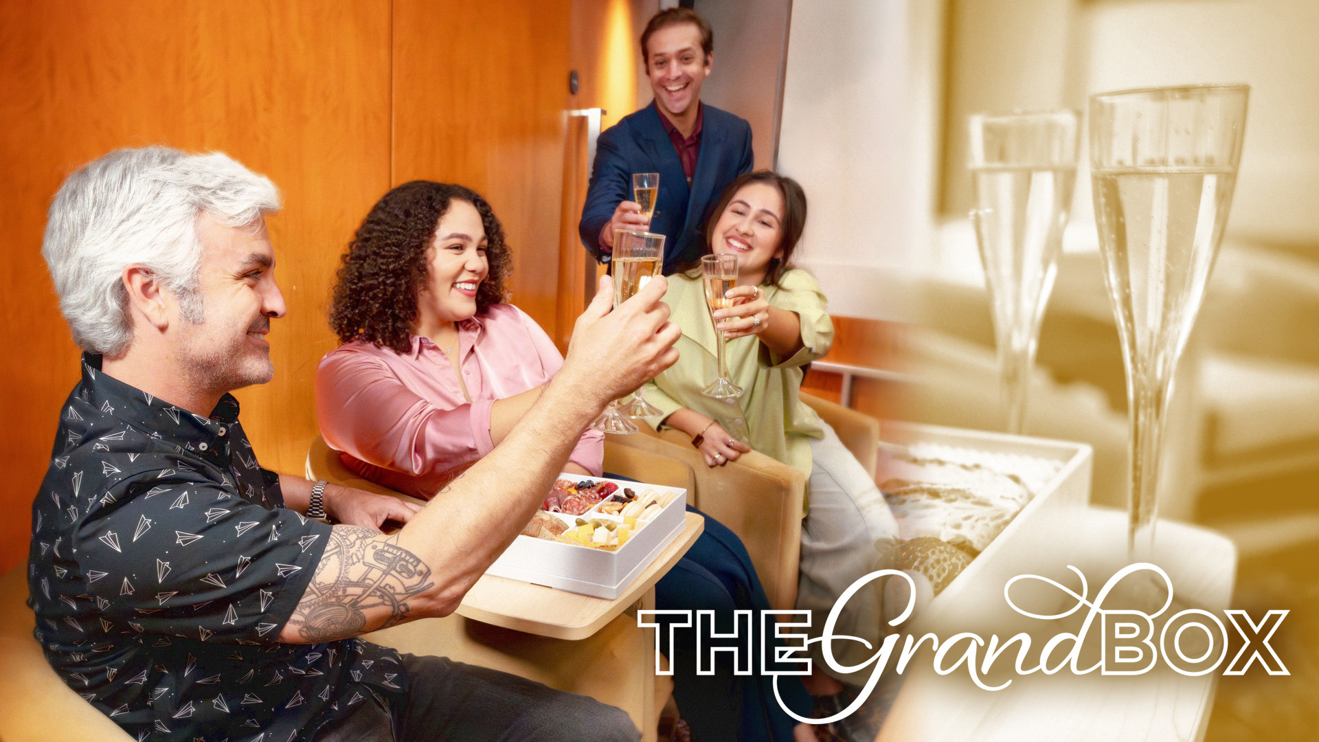Four adults in theatre box seats smiling and toasting champagne glasses, with a charcuterie box on the table.