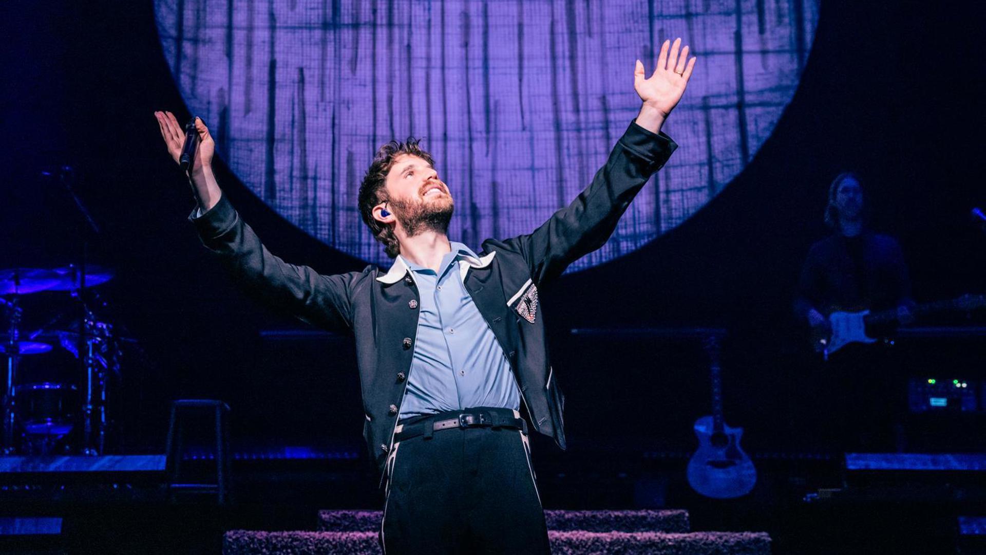 Ben Platt standing on stage with arms raised under blue and purple lighting, large circular backdrop behind.