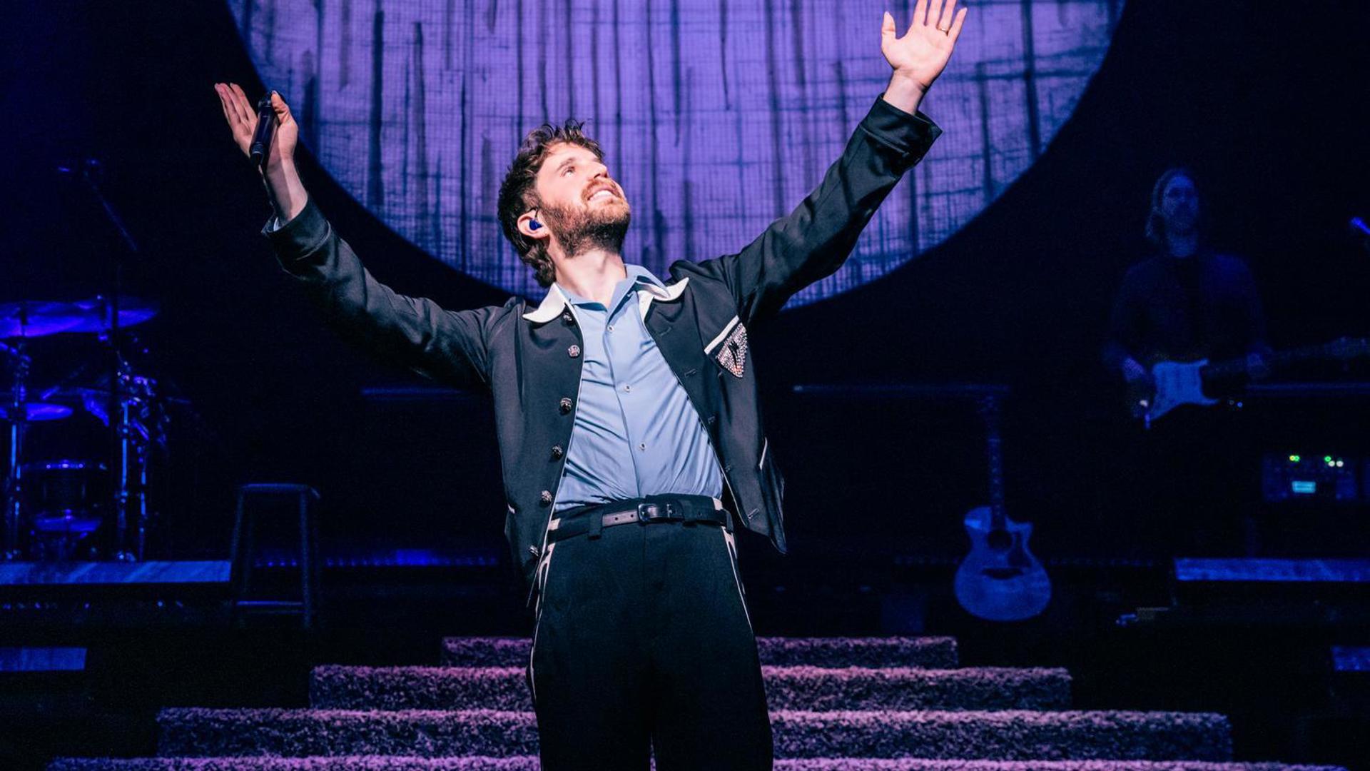 Ben Platt standing on stage with arms raised under blue and purple lighting, large circular backdrop behind.