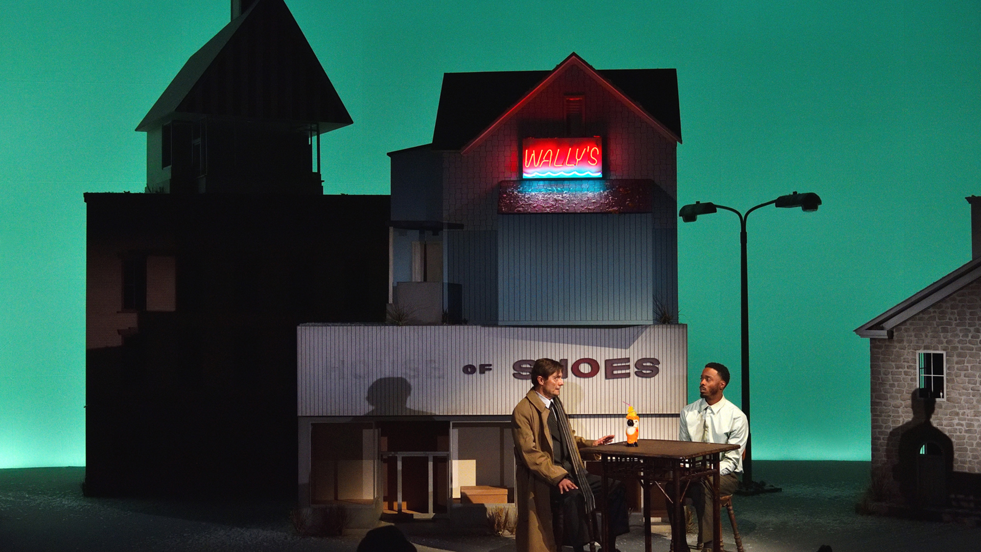Two men sit at a table with a tiki cocktail dink on it under a neon sign that says Wally's.