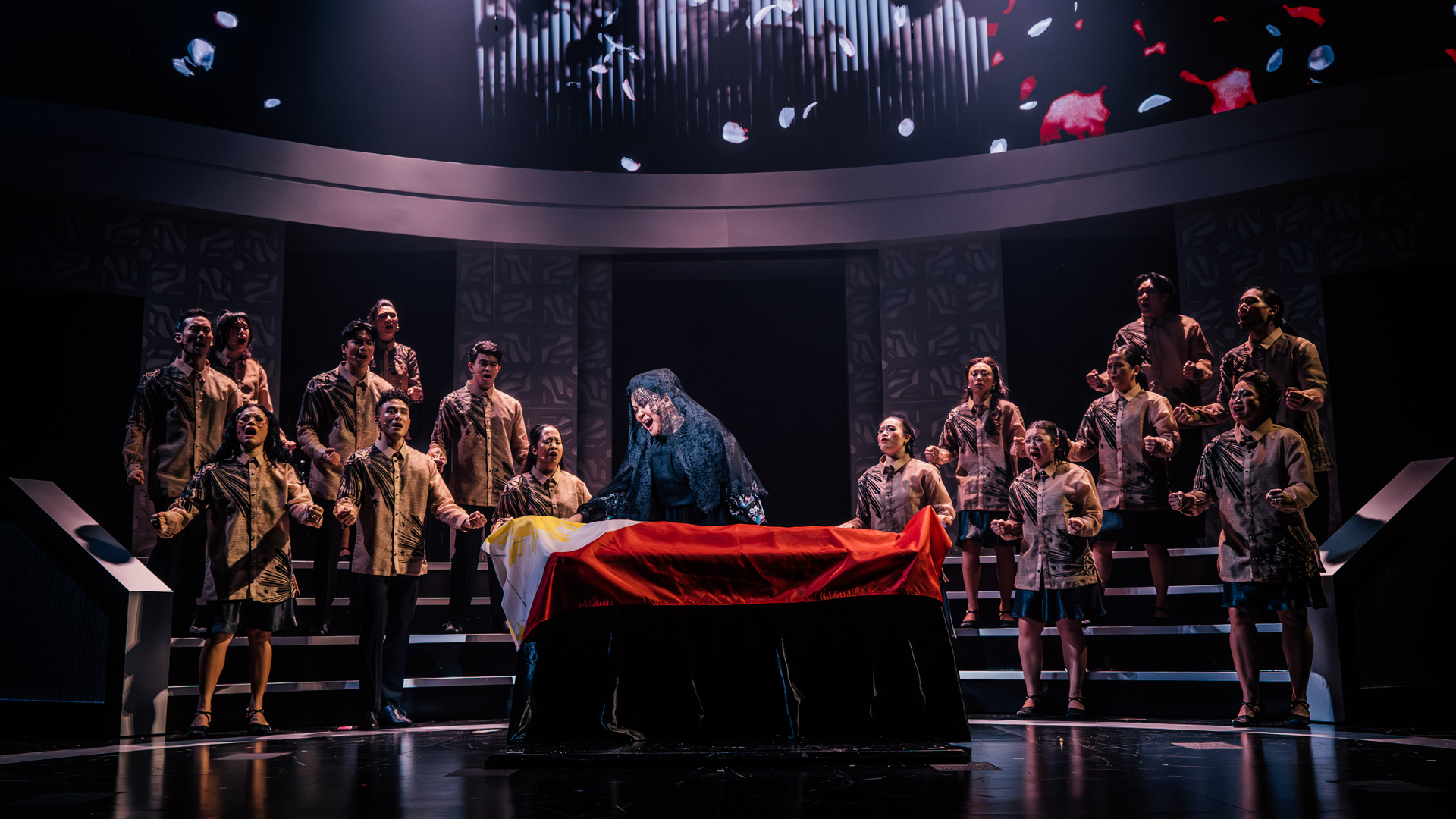 A performer caresses a casket draped in the Filipino flag. Performers stand on risers behind them, singing powerfully.