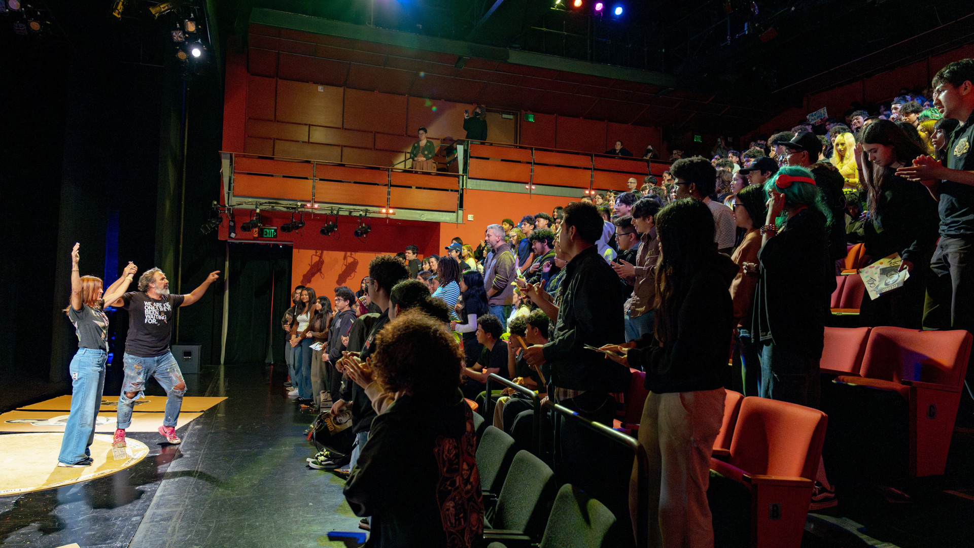 A lively audience stands and cheers during a performance in a theater.