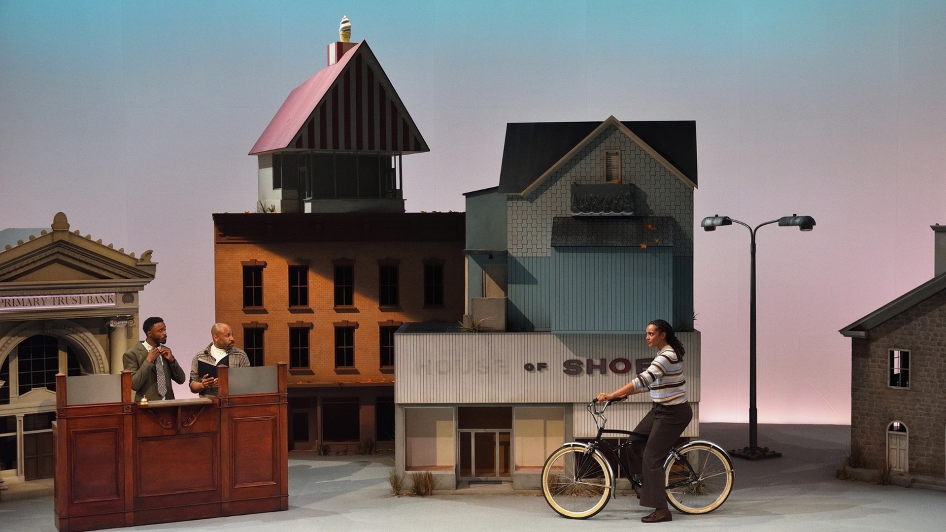 Two men stand behind the counter of a bank as a woman approached on a bicycle in a town full of large buildings.