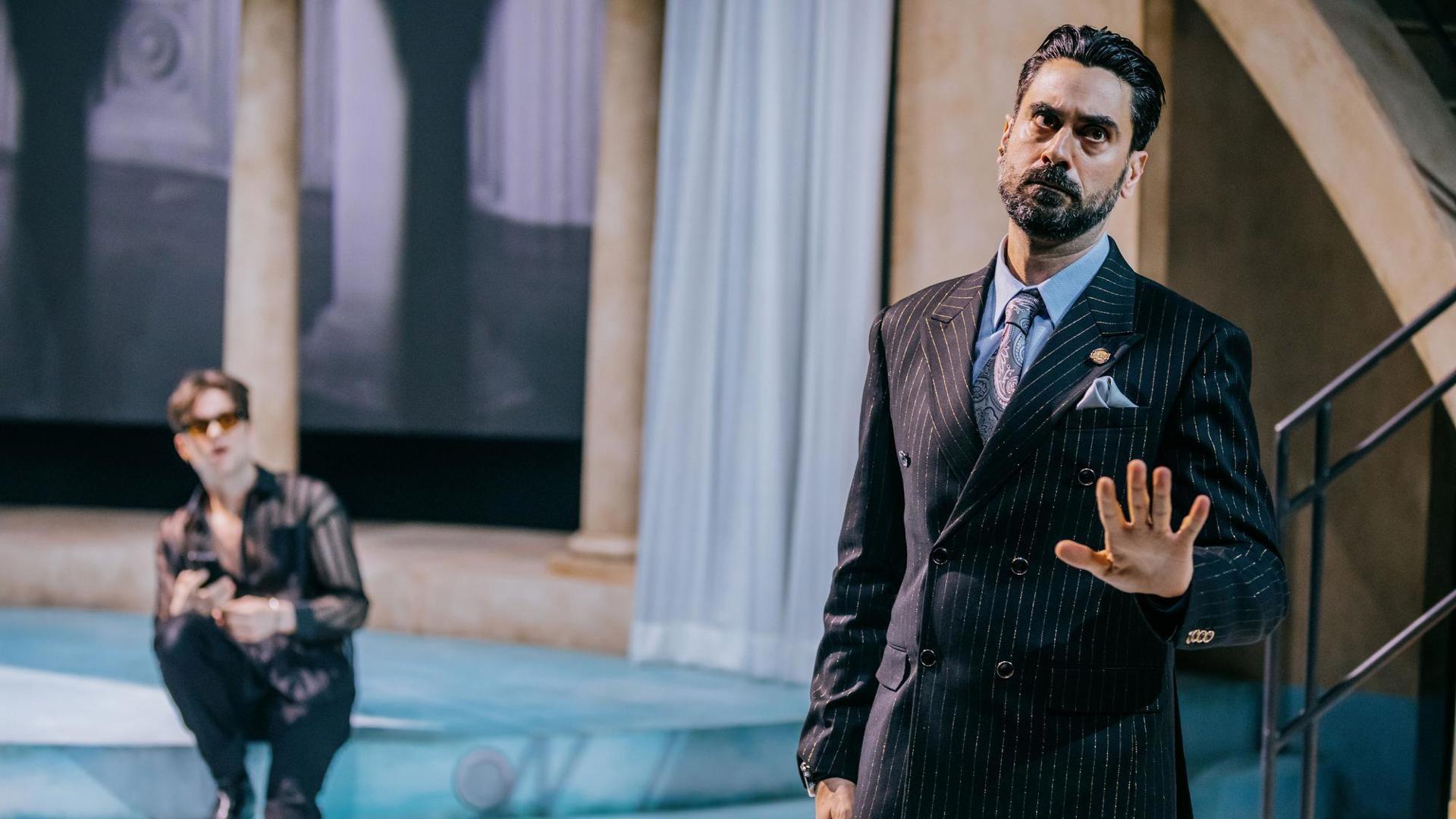 An actor stands downstage with their hand extended. Upstage, an actor watches with their mouth open.