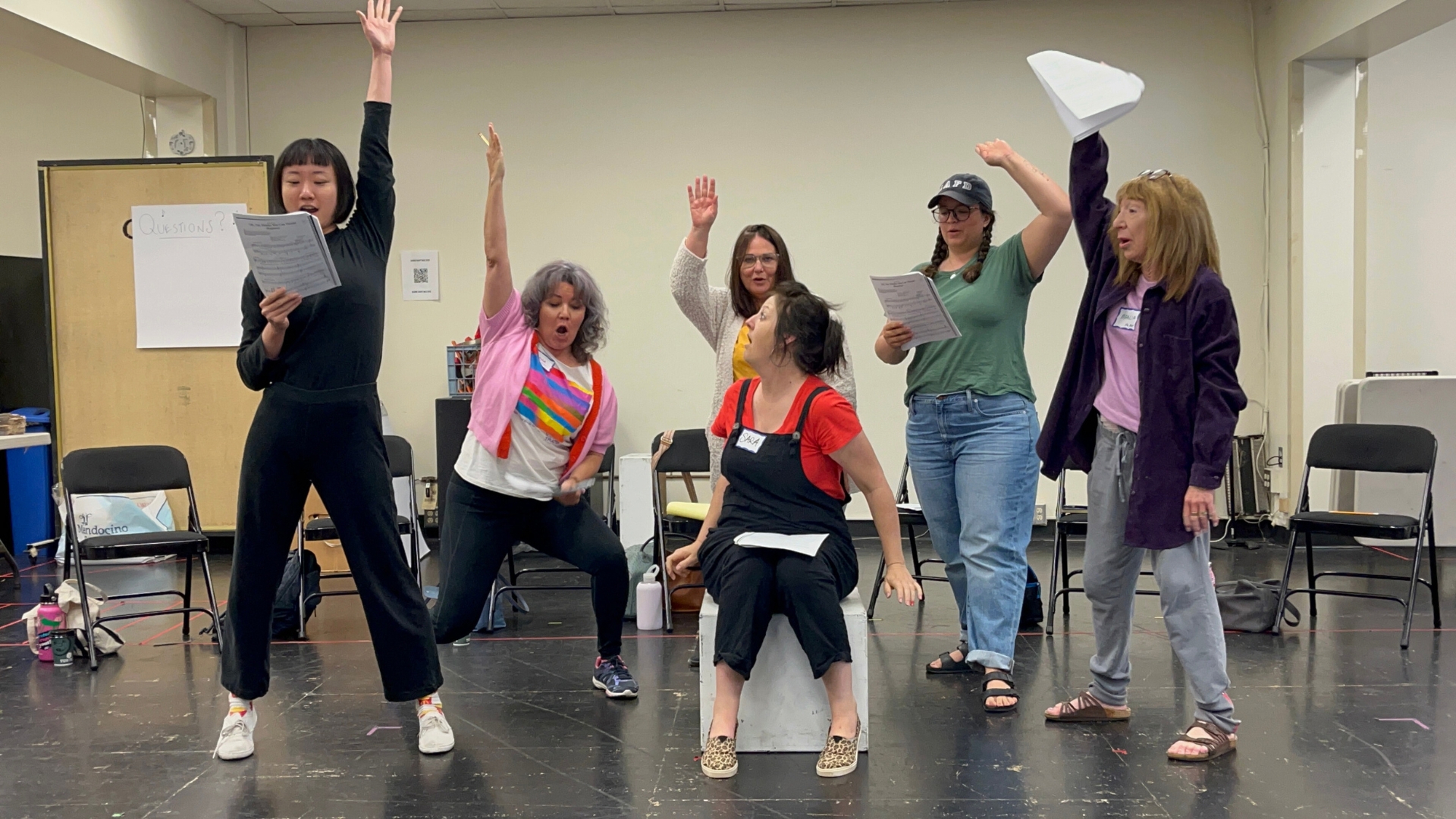 Group of six diverse educators in a rehearsal room, energetically performing with scripts in hand, raising their arms and striking playful poses.