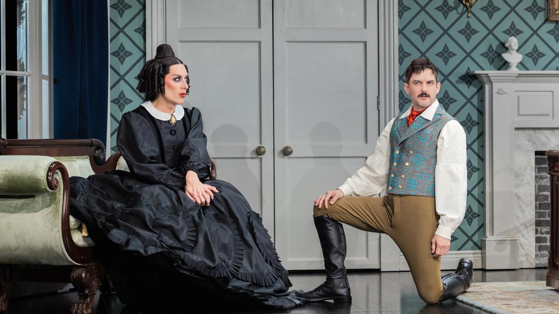 An actress sits on a green sofa in a black dress, an actor kneels to her right in black boots, brown pants and a blue vest, looking at the audience.