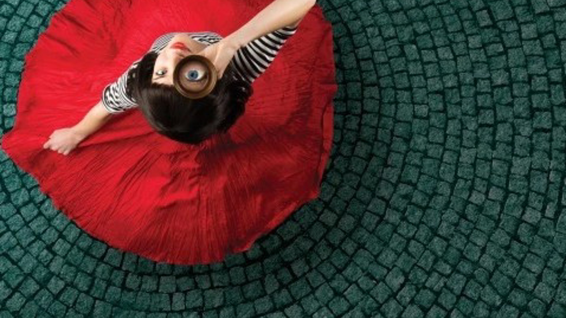 actress in red dress standing on a brick road looking up thru a scope for a new musical