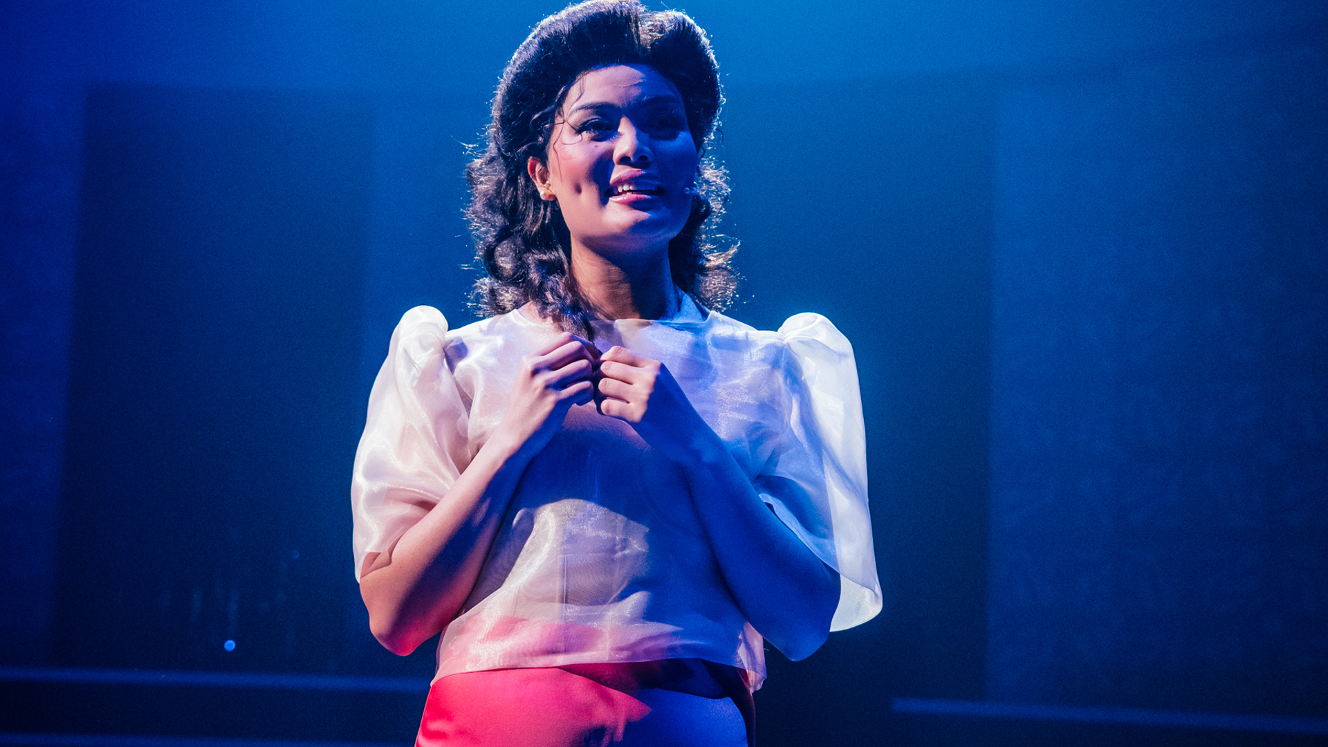 A performer stands, touching the ends of their hair while looking off dreamily with a smile. The words "IMELDA ROMUALDEZ, 16 YEARS OLD, TACLOBAN, PHILIPPINES" are projected onto the back wall.