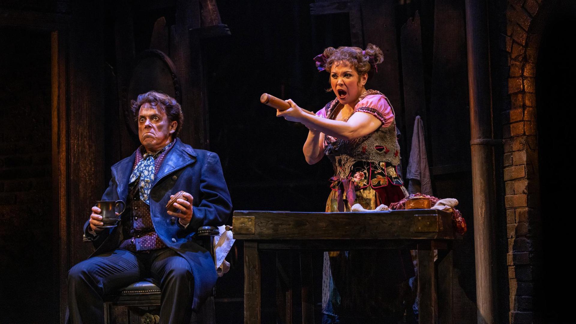 A performer sits, holding a cup and making a disgusted face. Another performer stands next to him, singing intensely and holding up a rolling pin like a weapon.