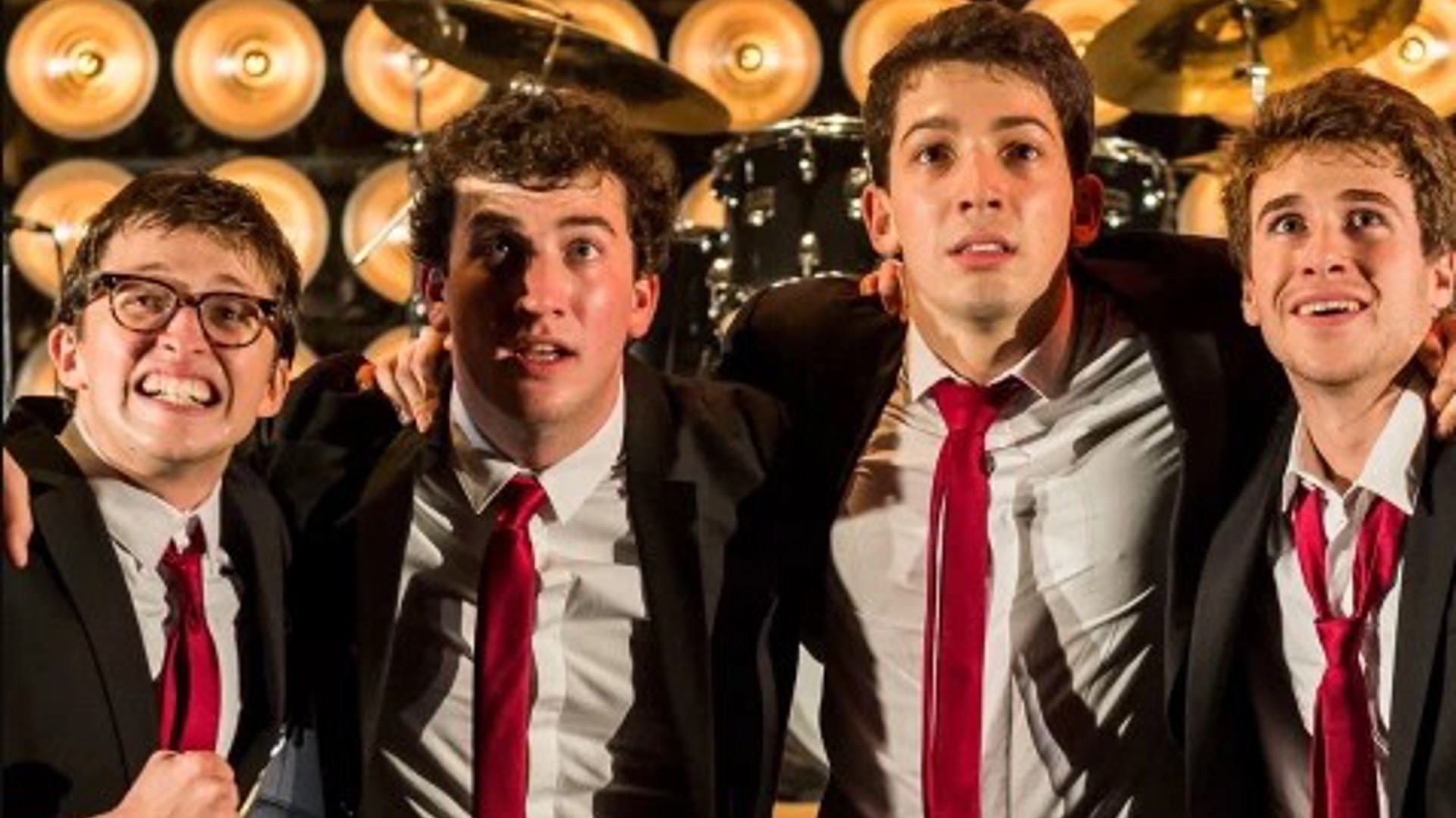 four wide eyed young men arm in arm in suits in front of a drum kit
