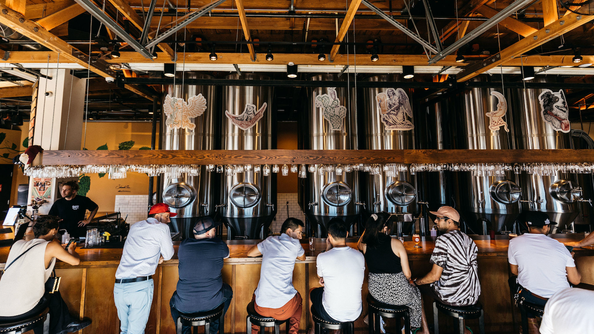 Patrons sit at bar conversing. Beer steel vats are in the background. 