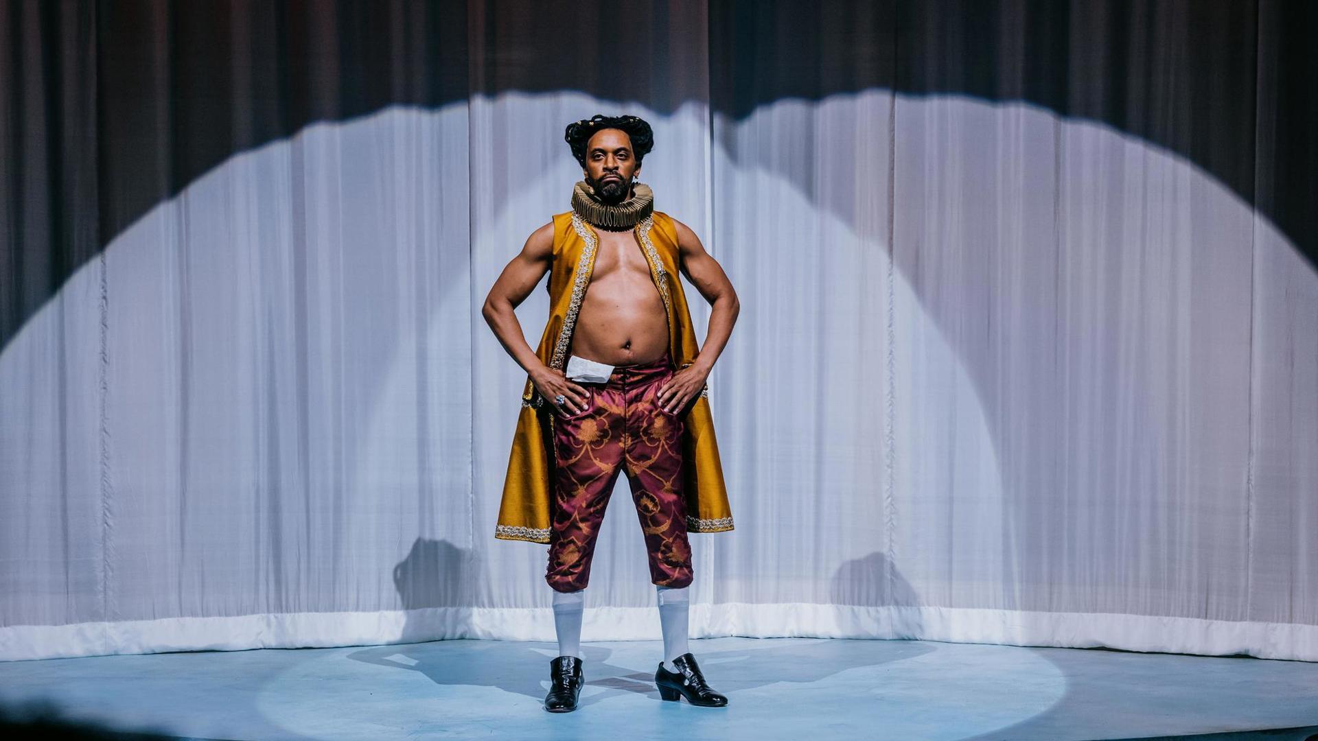An actor stands akimbo center stage, wearing flamboyant Shakespearean clothing.