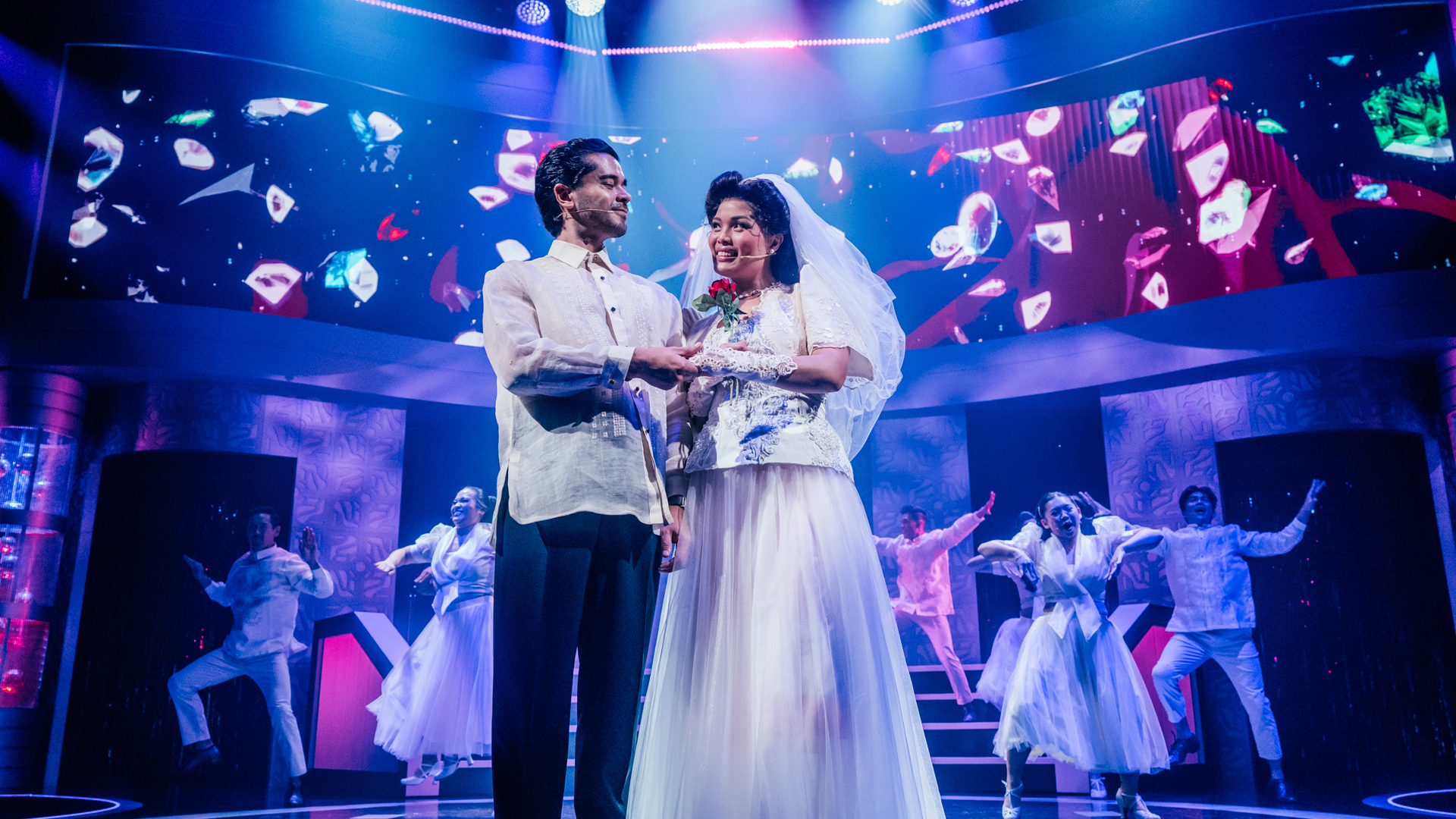 Two performers stand at the edge of the stage, getting married as dancers dance behind them.
