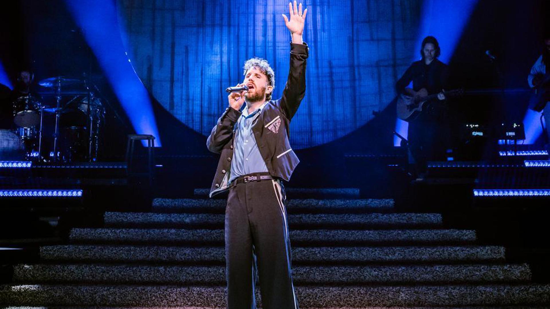 Ben Platt singing on stage with one arm raised under bright blue spotlights and circular backdrop.