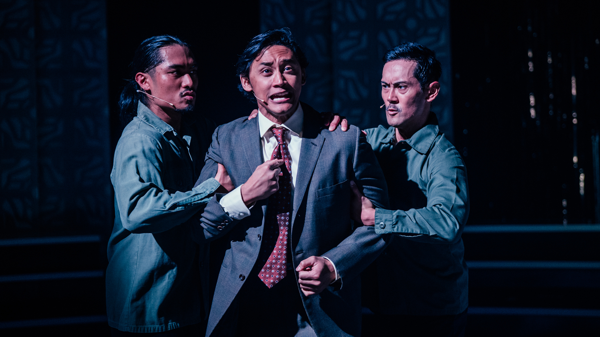 A performer has an anguished expression as they are aggressively held back by the arms by two ensemble members.