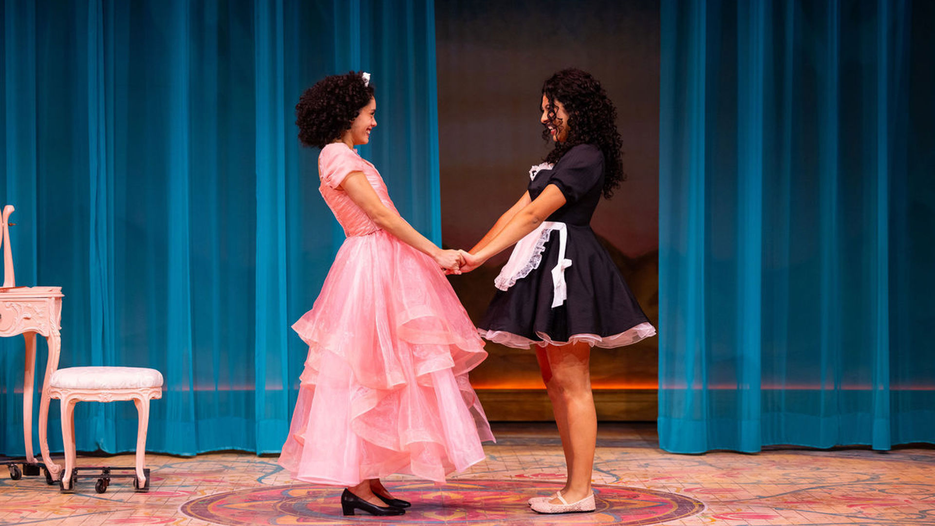 Two actors, one in a large pink dress and the other in a maid uniform hold hands and smile at each other.