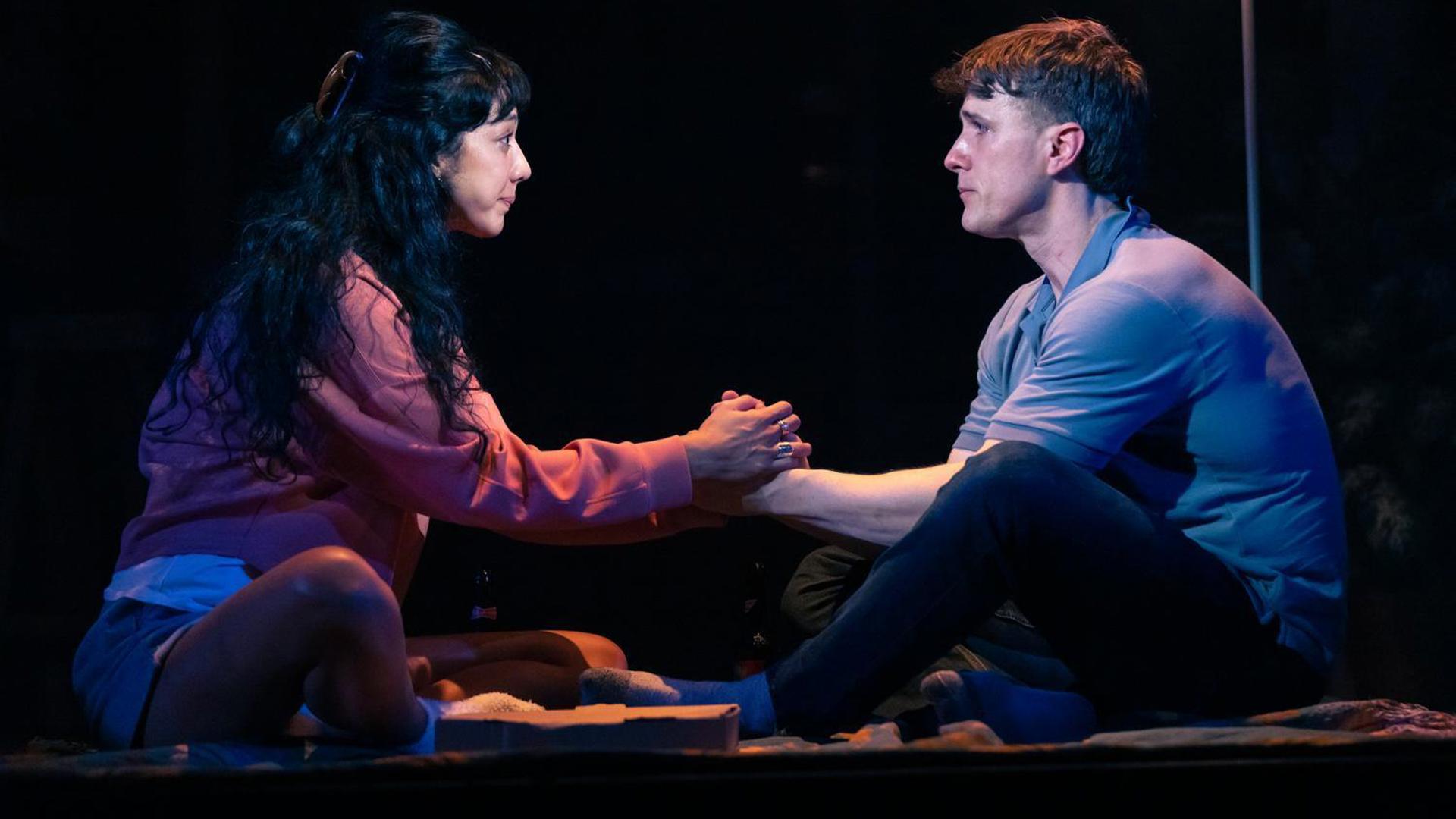 A couple holds hands while sitting on the floor. Their faces are distraught.