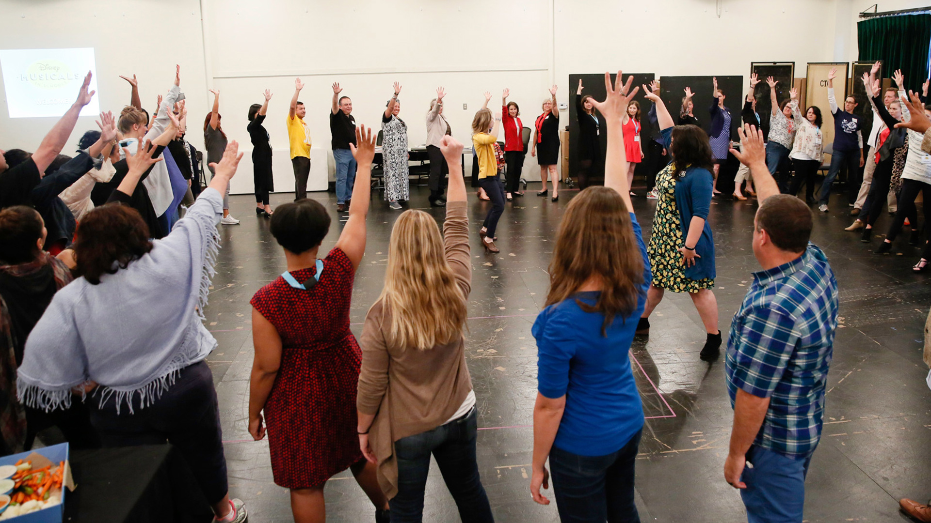 A large group of people raise their hand and form a circle within the rehearsal room.