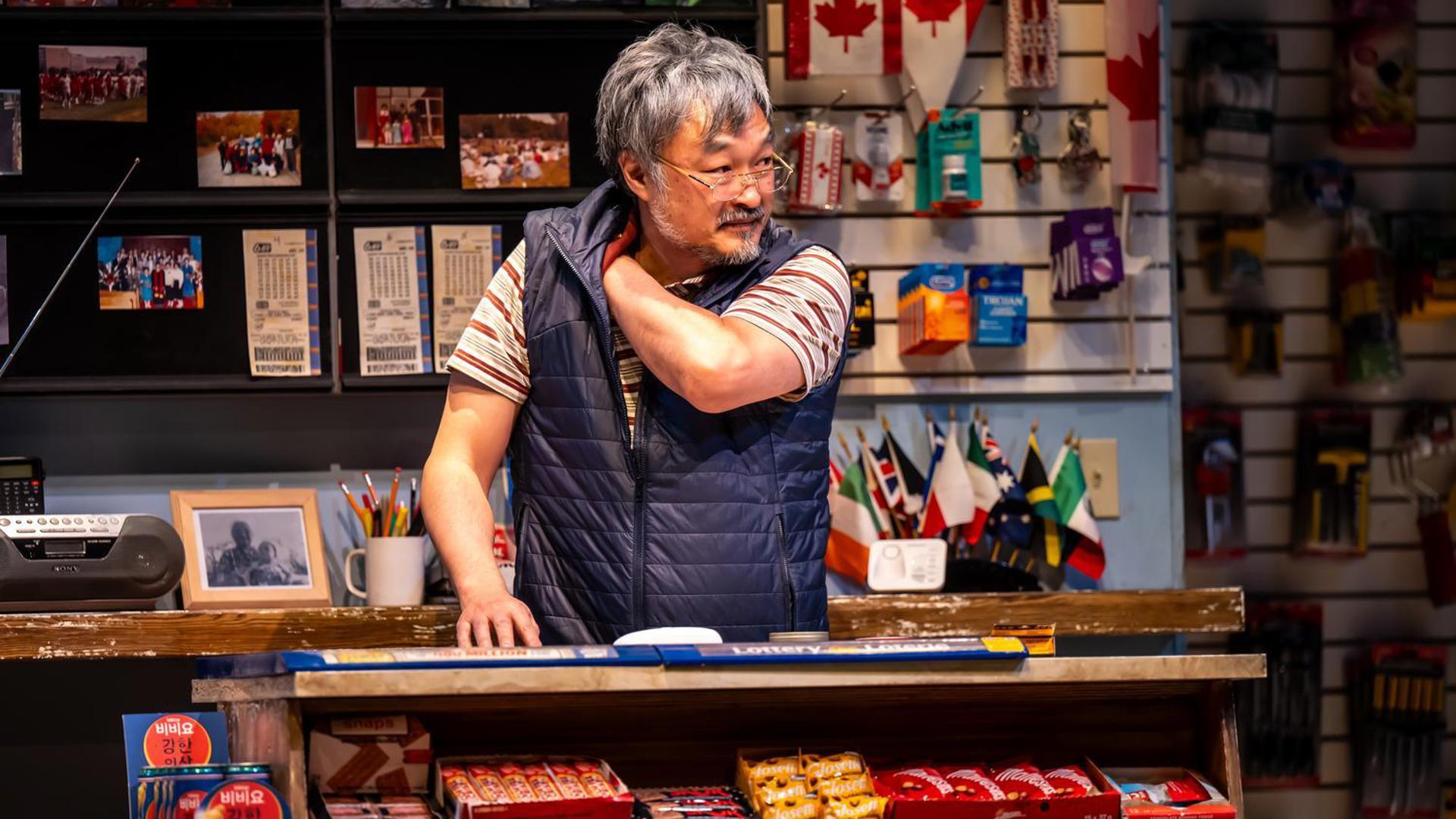 A man stands behind a counter scratching his shoulder. 