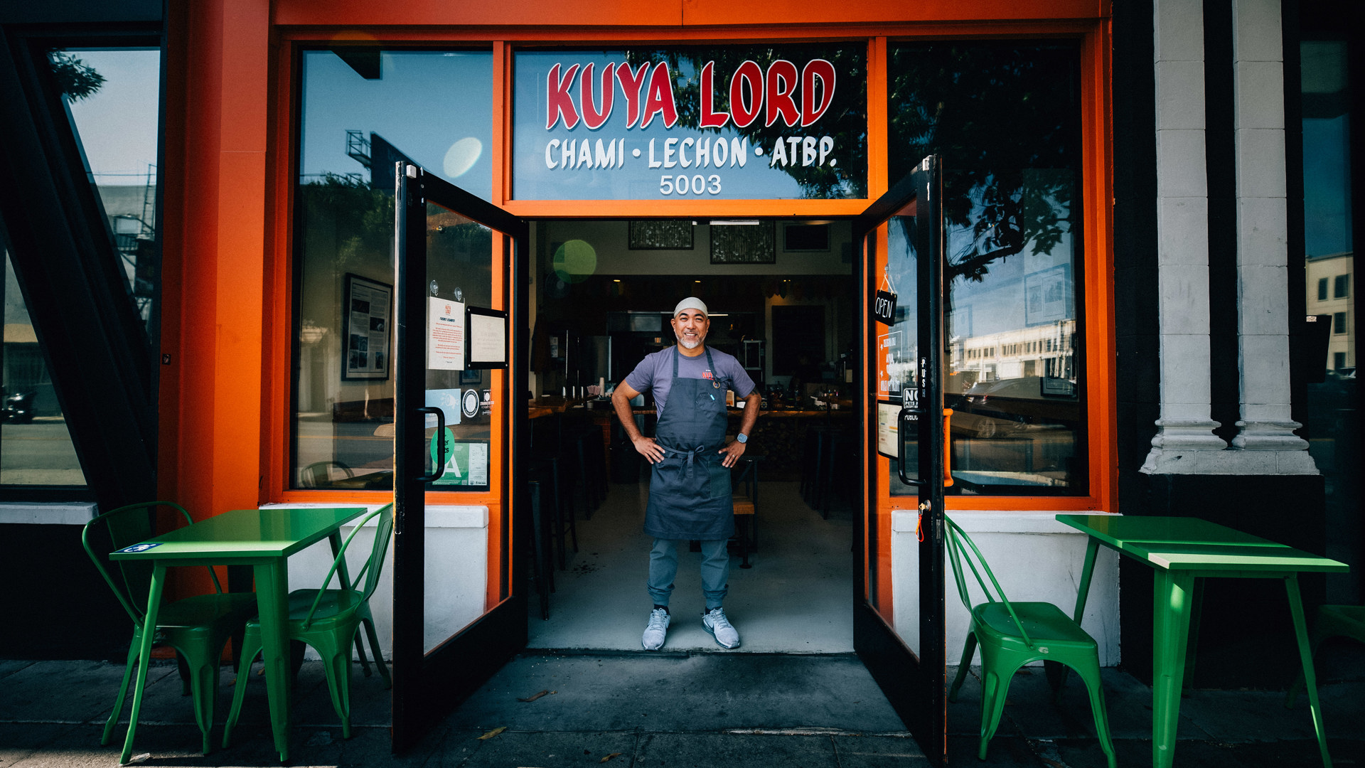 A man in an apron stands smiling in the doorway of a restaurant called “Kuya Lord.”