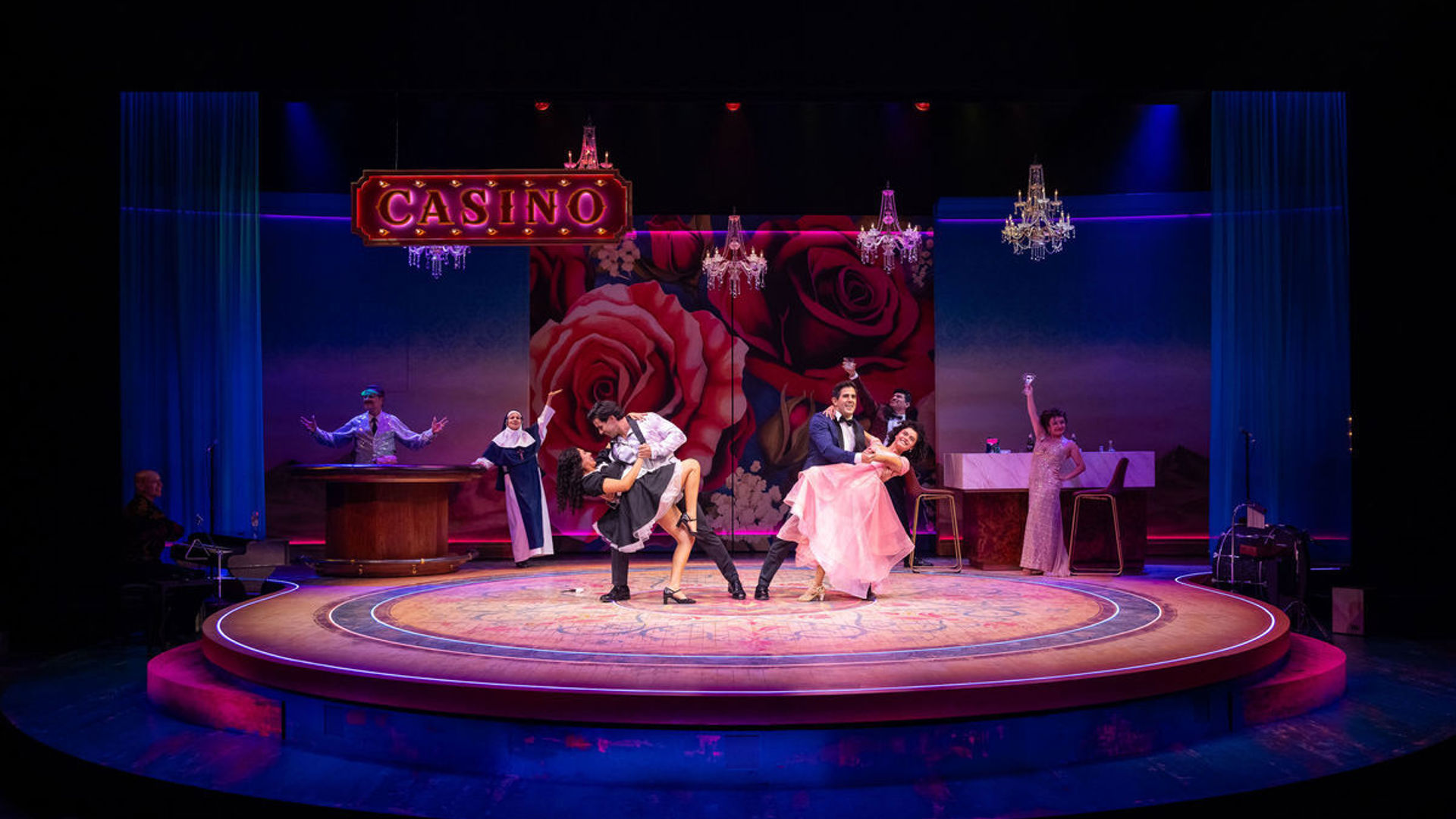 A group of actors pose onstage under a sign that reads 'CASINO'. Center stage, two couples dip in a dance move.