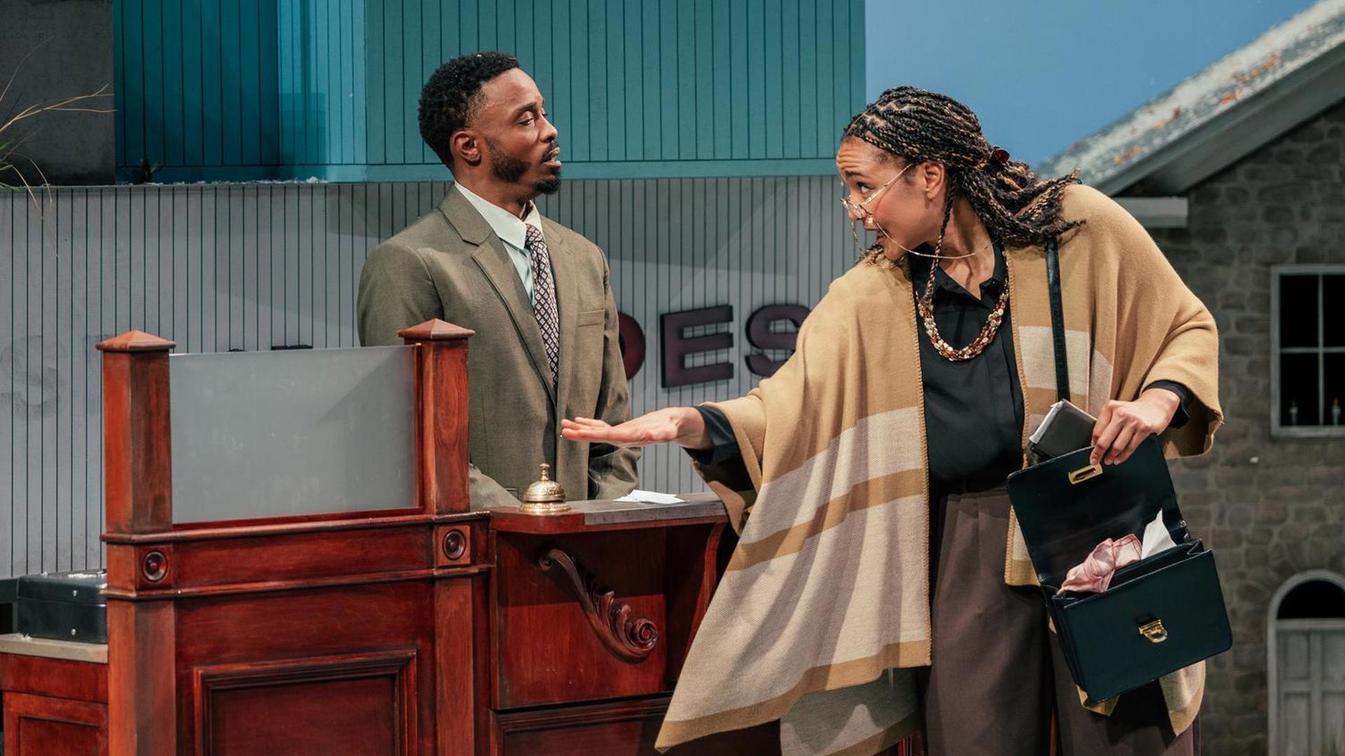 A man in a suit stands behind a bank counter, while a woman with glasses, a purse, and a shawl rings look at the man while ringing a desk bell.