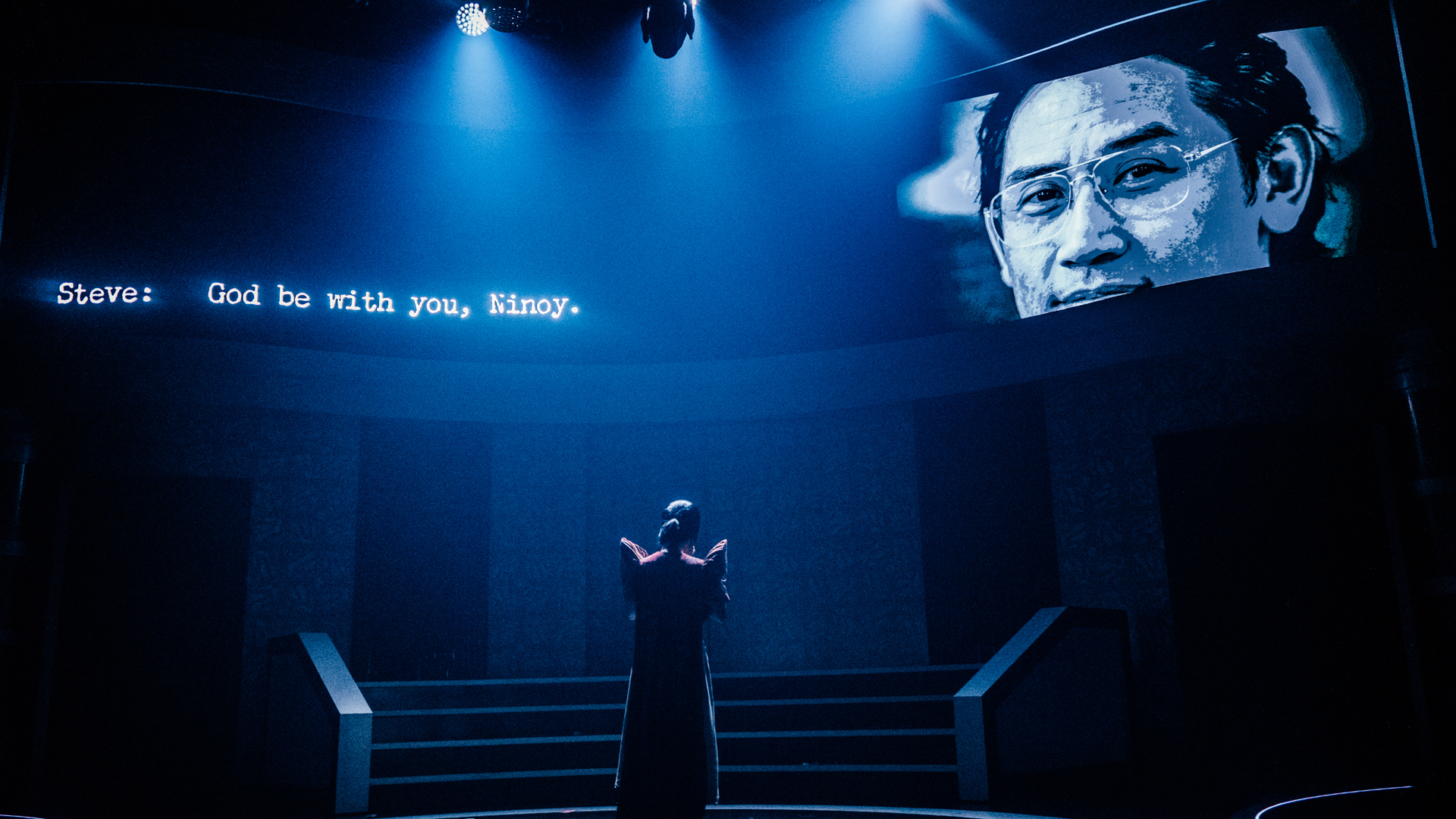 A performer stands center stage, facing the back wall of the stage. A picture of a man, and the words "Steve: God be with you, Ninoy" are projected onto the back wall.