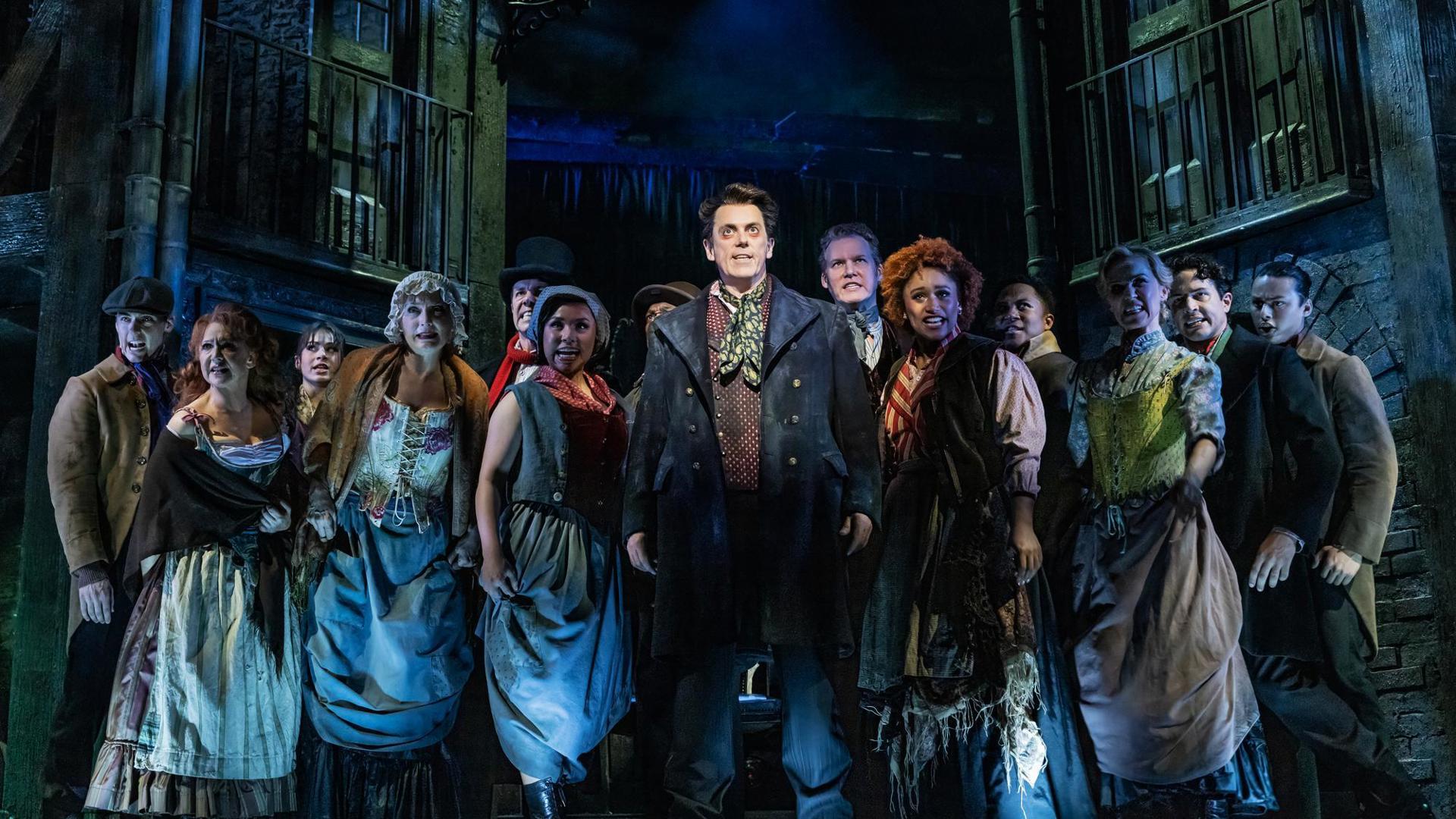 Ensemble members stand in a tight clump onstage. The lighting is dark and dramatic.