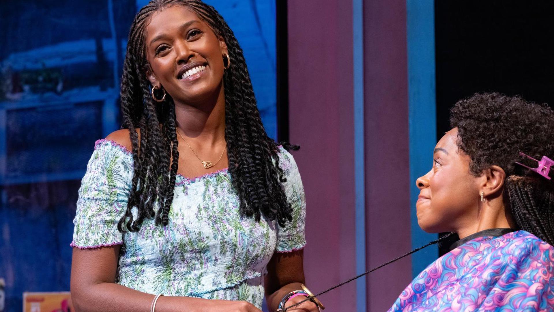 A performer looks off into the distance with a smile while braiding another performer's hair.