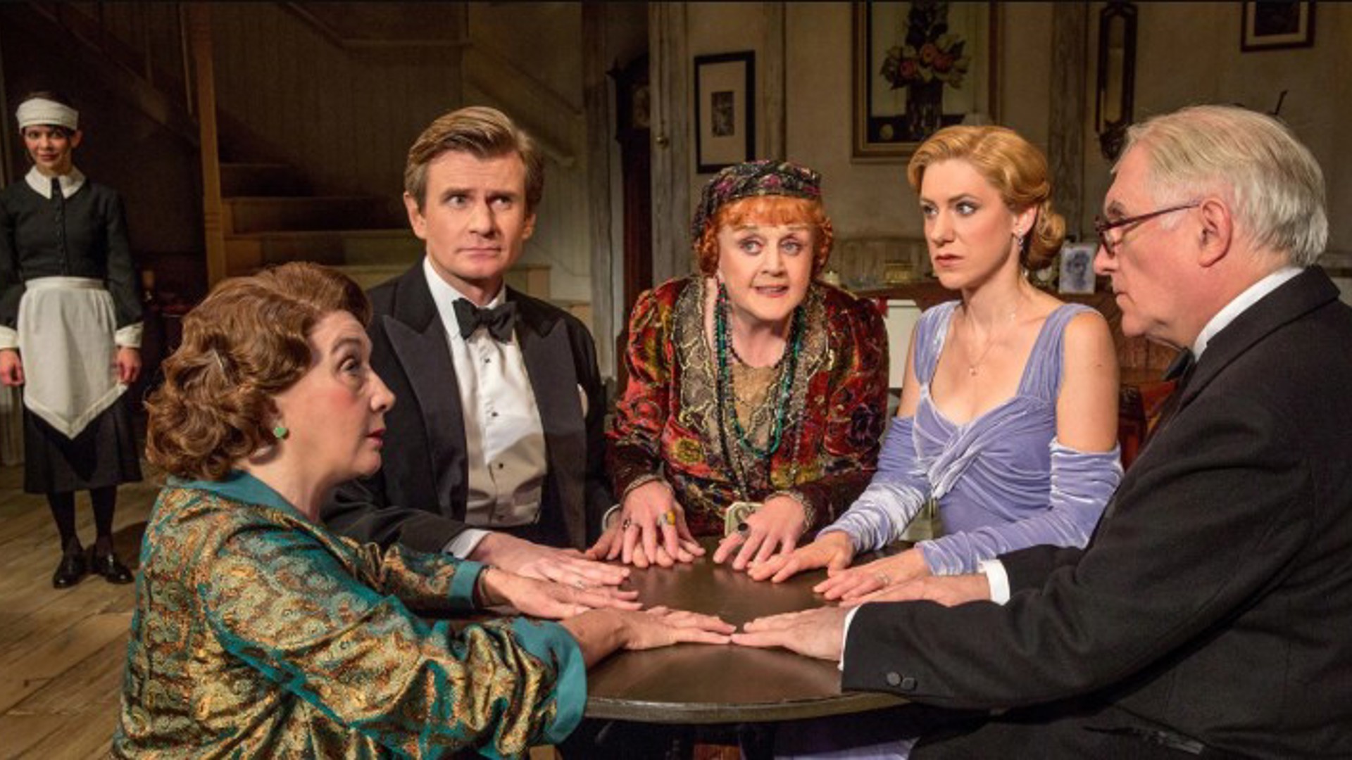 the cast of Blithe Spirit preforming around a séance table with a maid standing in the background