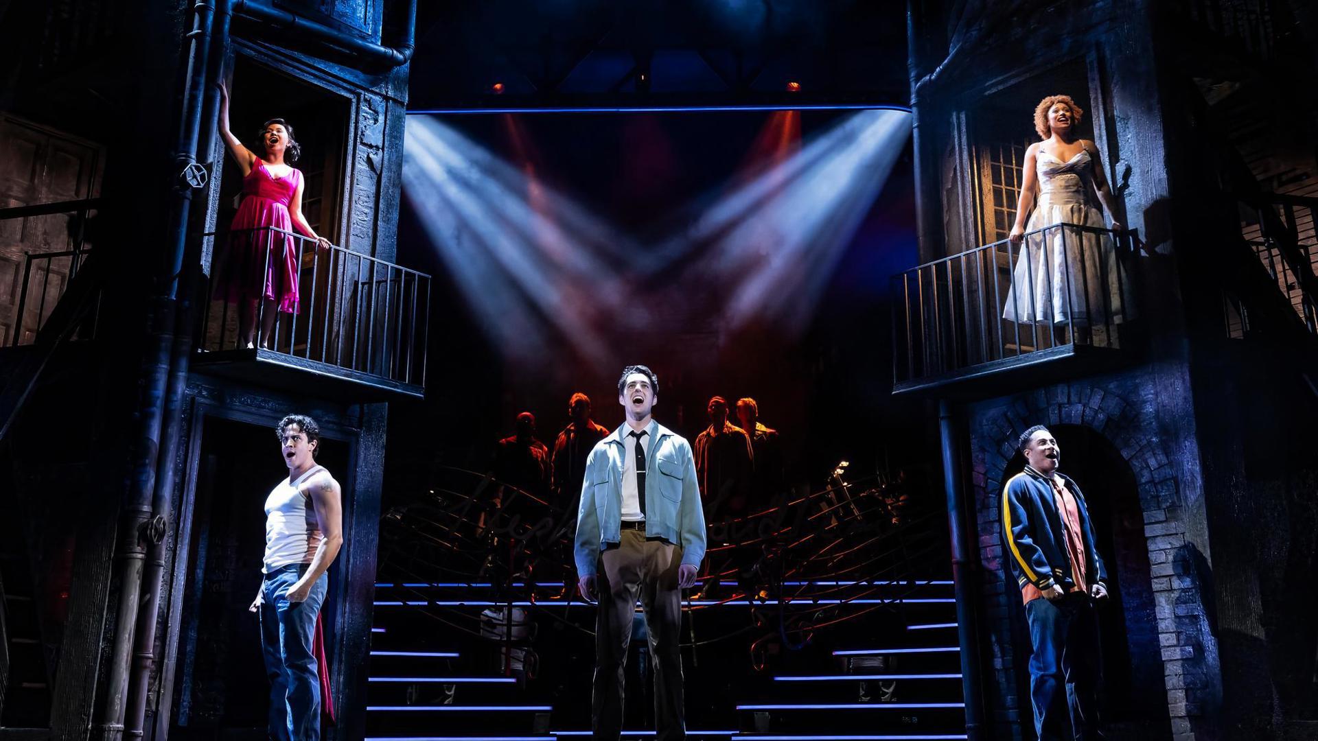 Five performers sing out toward the audience. Three stand on the floor and two stand on balcony set pieces.