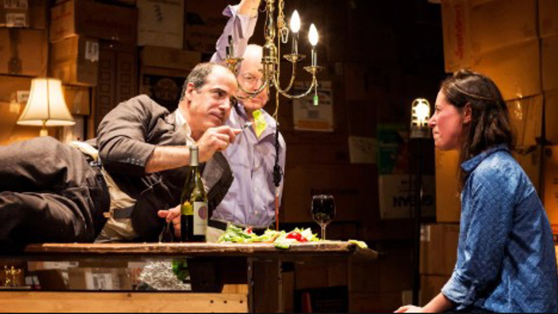 Geoff Sobelle lying sideways on a table with an actor behind and an actress sitting in front