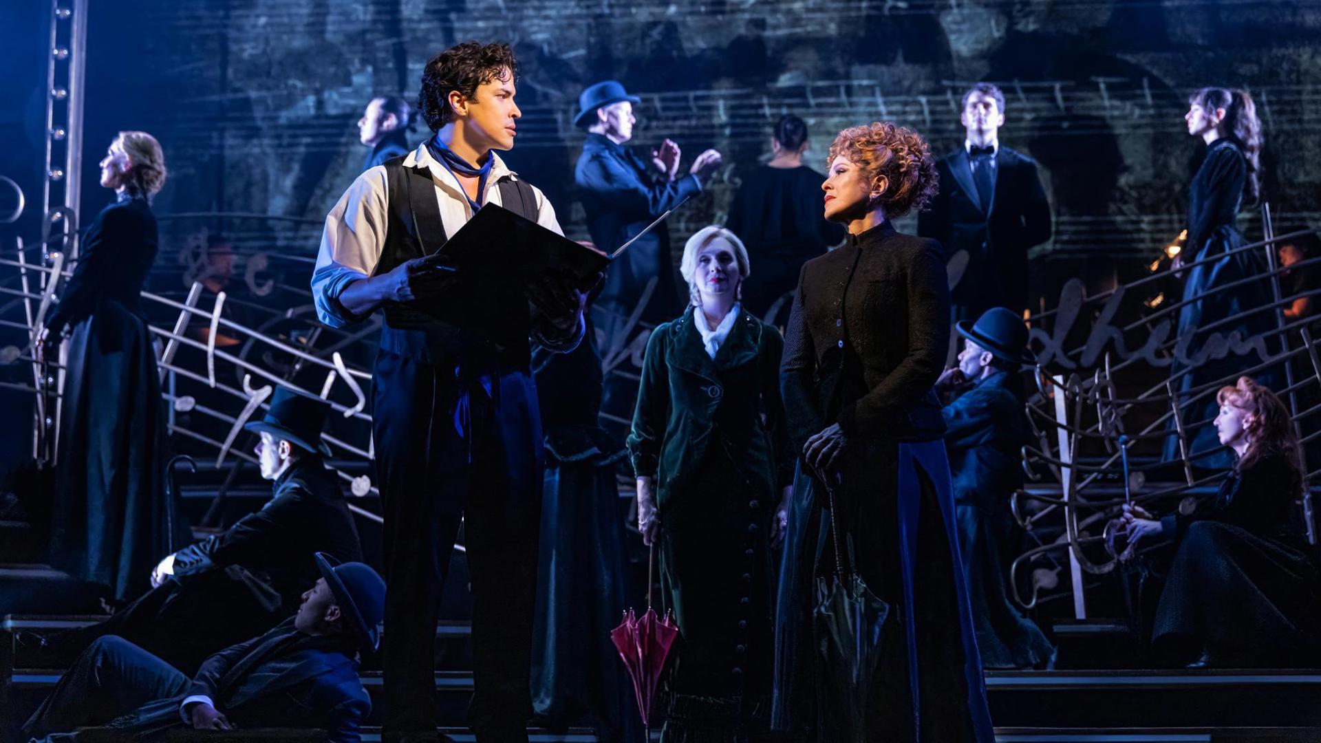 Two performers stand center stage, looking at each other. One holds a book, the other holds a parasol. In the background, ensemble members stand and sit facing different directions.