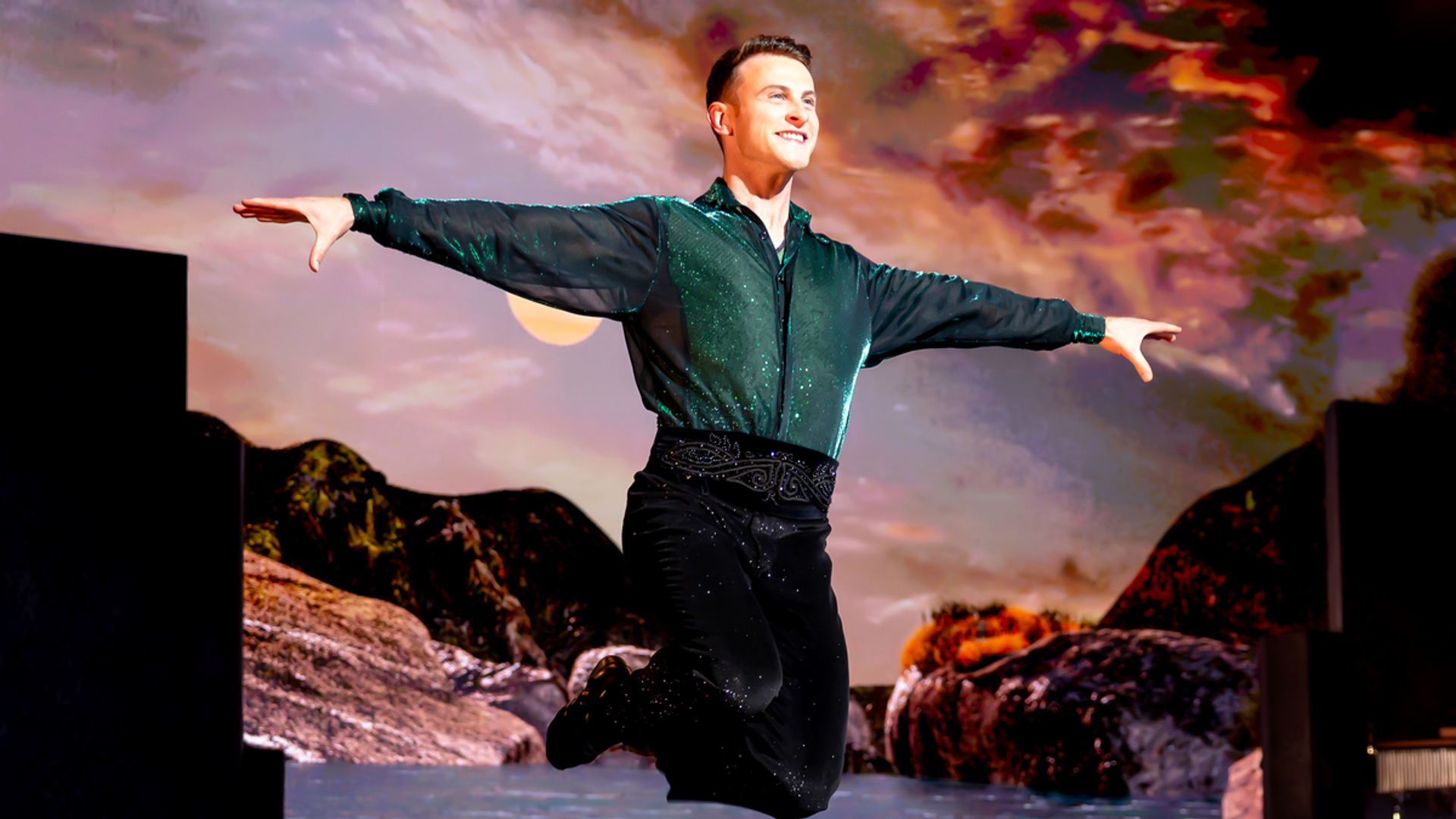 A performer jumping on stage with arms extended and legs behind him.