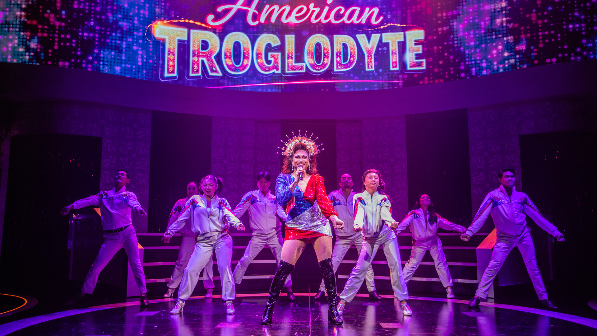 A drag performer stands center stage singing into a microphone while dancers in vintage track suits dance behind them. The words "American Troglodyte" are projected onto the back wall.