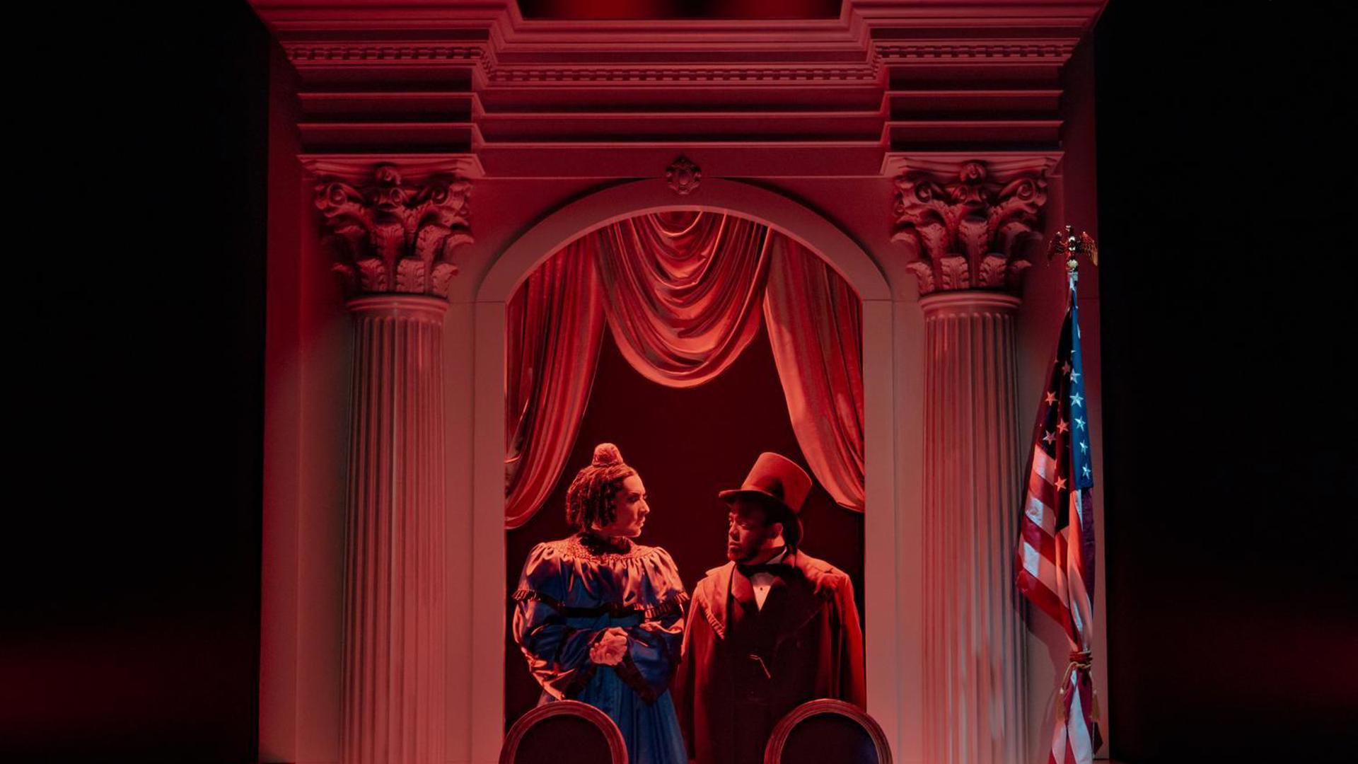 Two actors face each other standing in a theater box. There is an American flag to their right and the box is washed in red light.