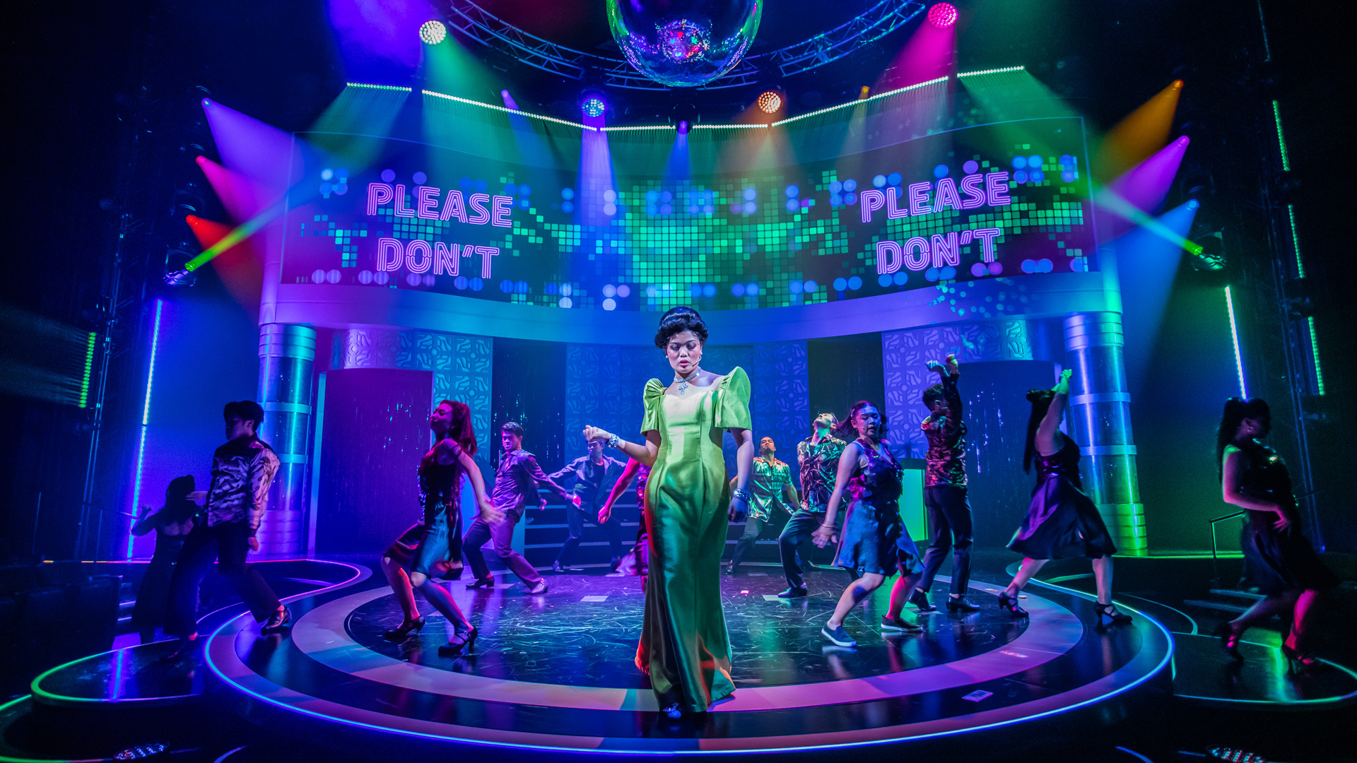 A performer stands center stage beneath a large disco ball while dancers, lit by multicolored lights, dance in the background. The words "Please Don't" are projected onto the back wall.