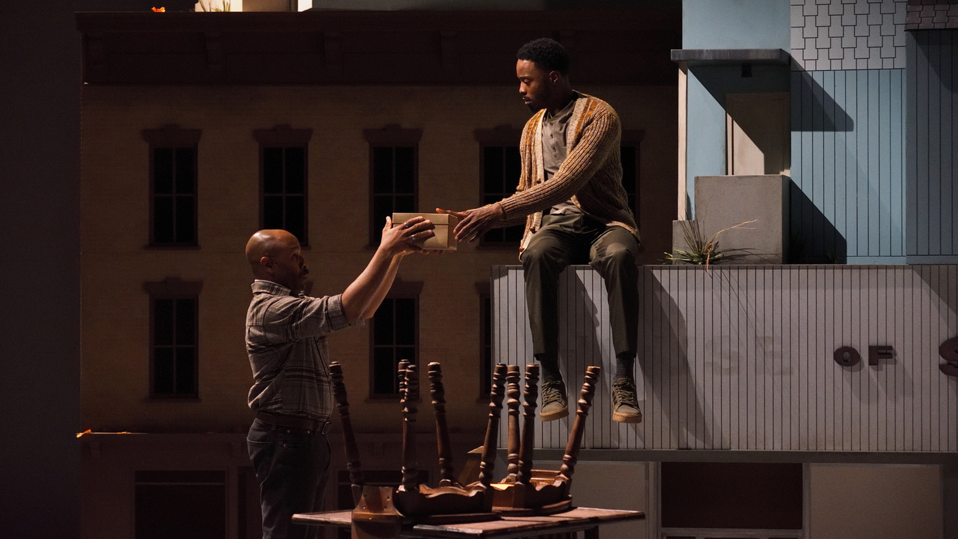 A man hands another man a shoebox in front of a table with chairs on top it.