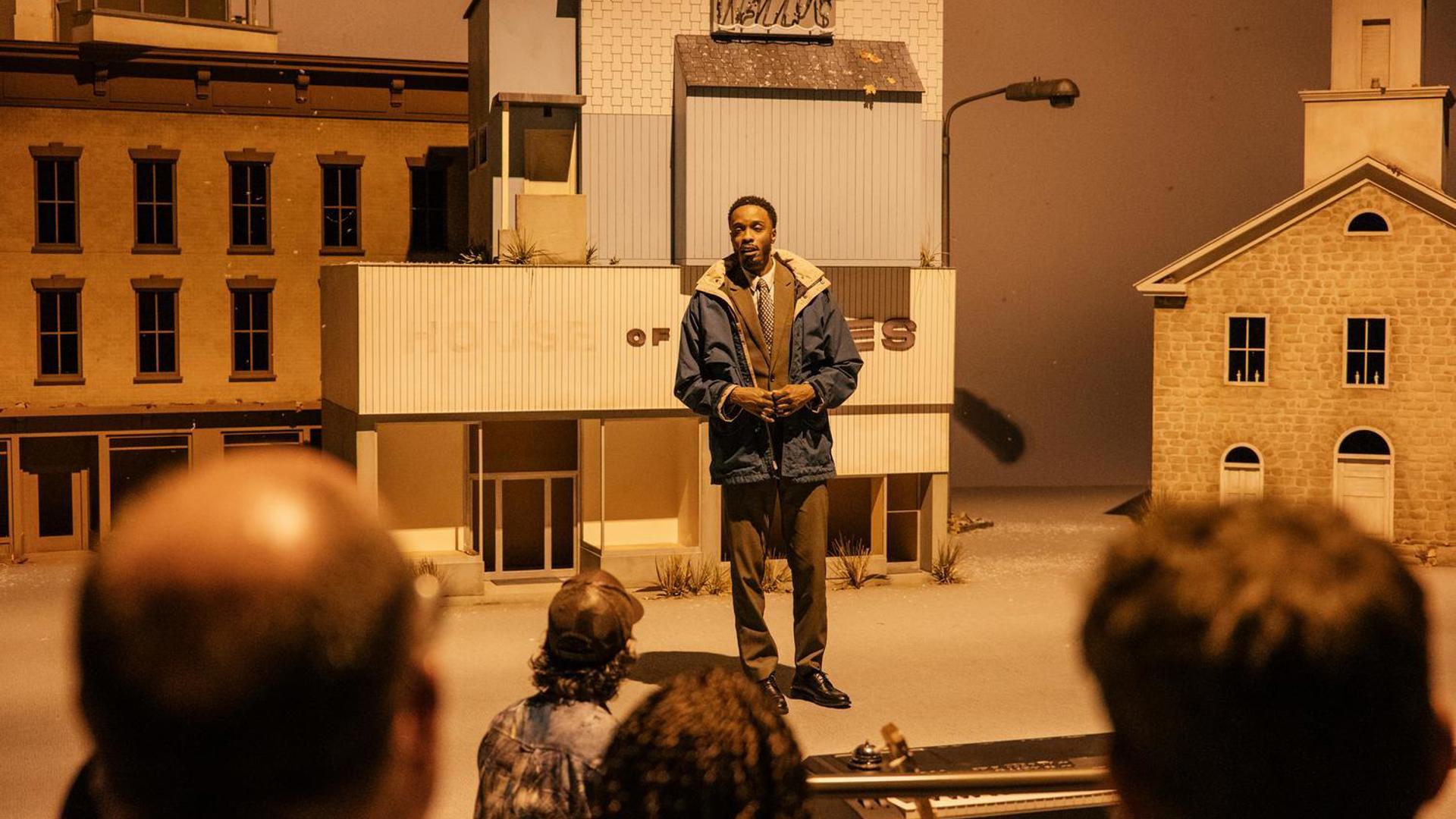 A man in a jacket, suit, and tie stands on stage in front of large buildings looking out at the audience.