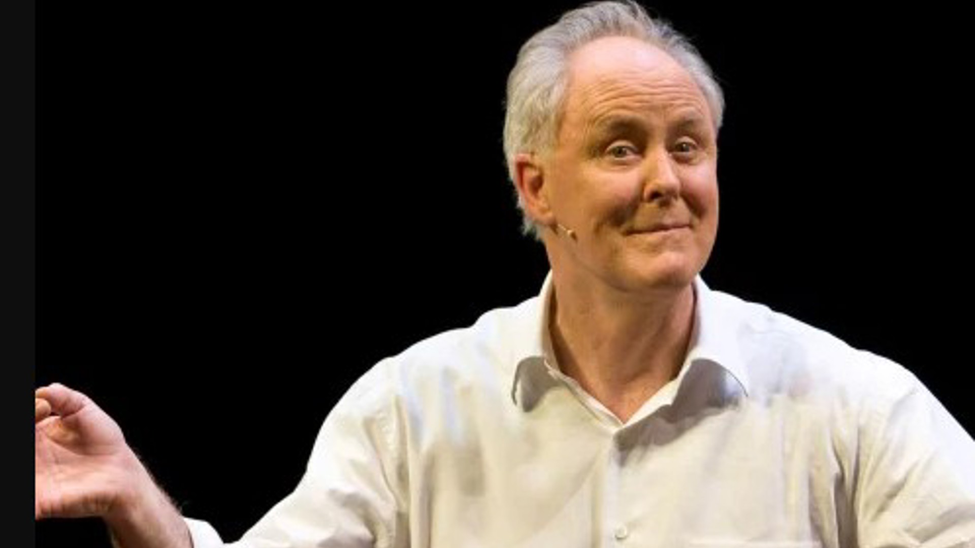 John Lithgow standing with arms open, grinning, with a small headset microphone on