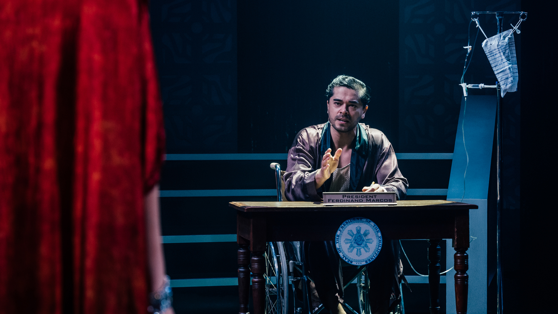 A performer sits  at a desk in a wheelchair, addressing an unseen person in front of them. They are rigged up to an IV drip and a plaque on the desk reads "President Ferdinand Marcos".