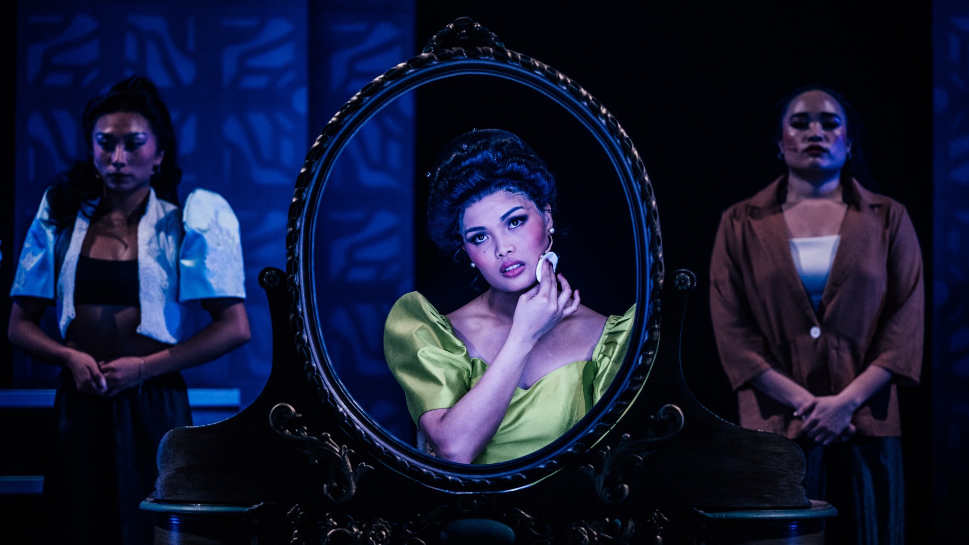 A performer sits at a mirror, applying makeup. Ensemble members stand in the background with their hands folded in front of themselves.
