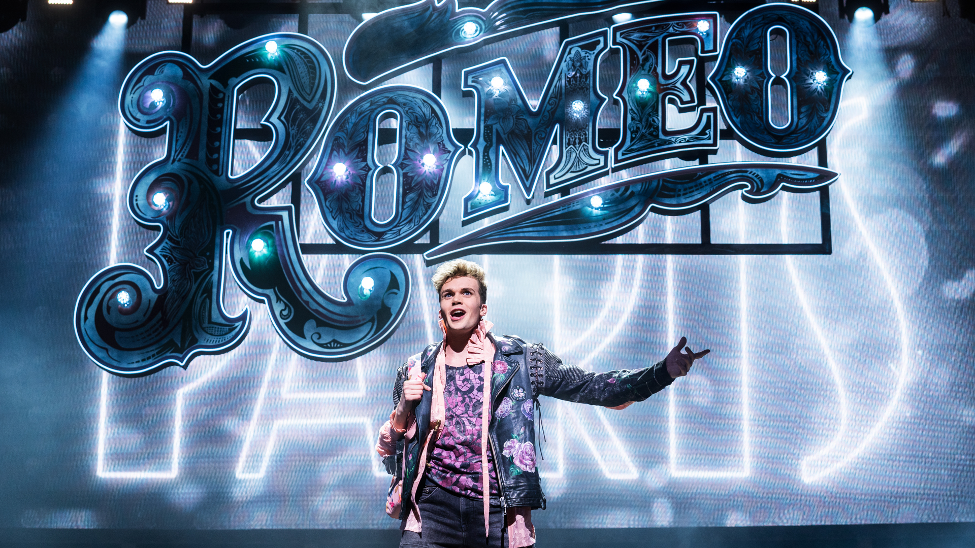 An actor stands center stage with one arm out. Giant LED sign displays ROMEO/PARIS behind the actor.