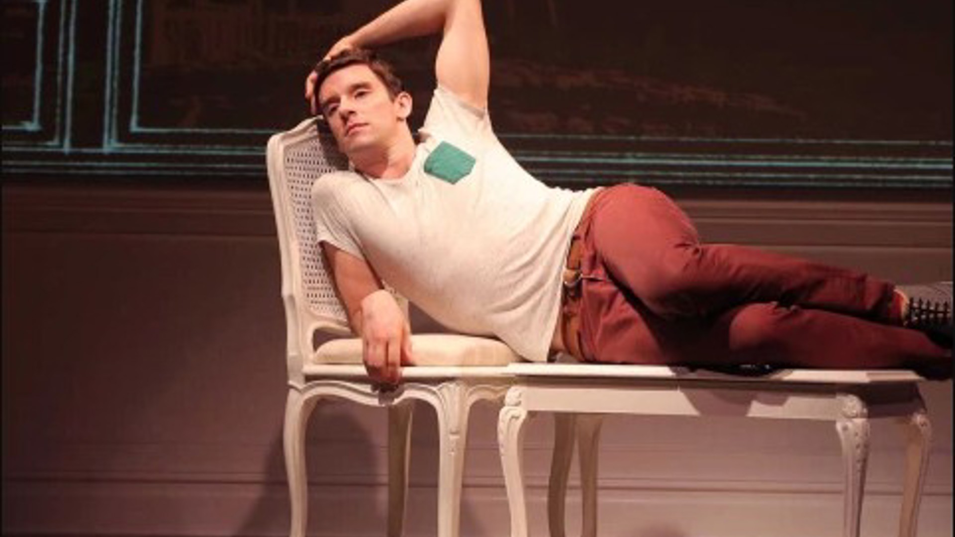 Michael Urie lying across a table and chair on stage at the Mark Taper Forum
