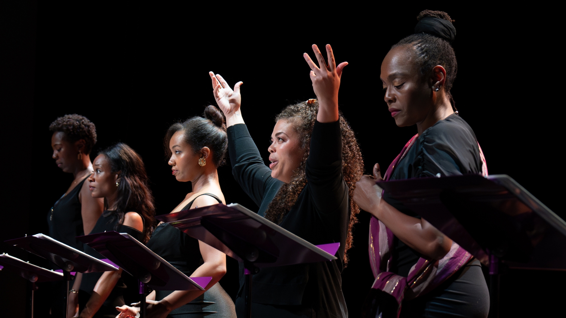 A group of performers stand in line infront of music stands, the center performer holds both of their arts above their heads.