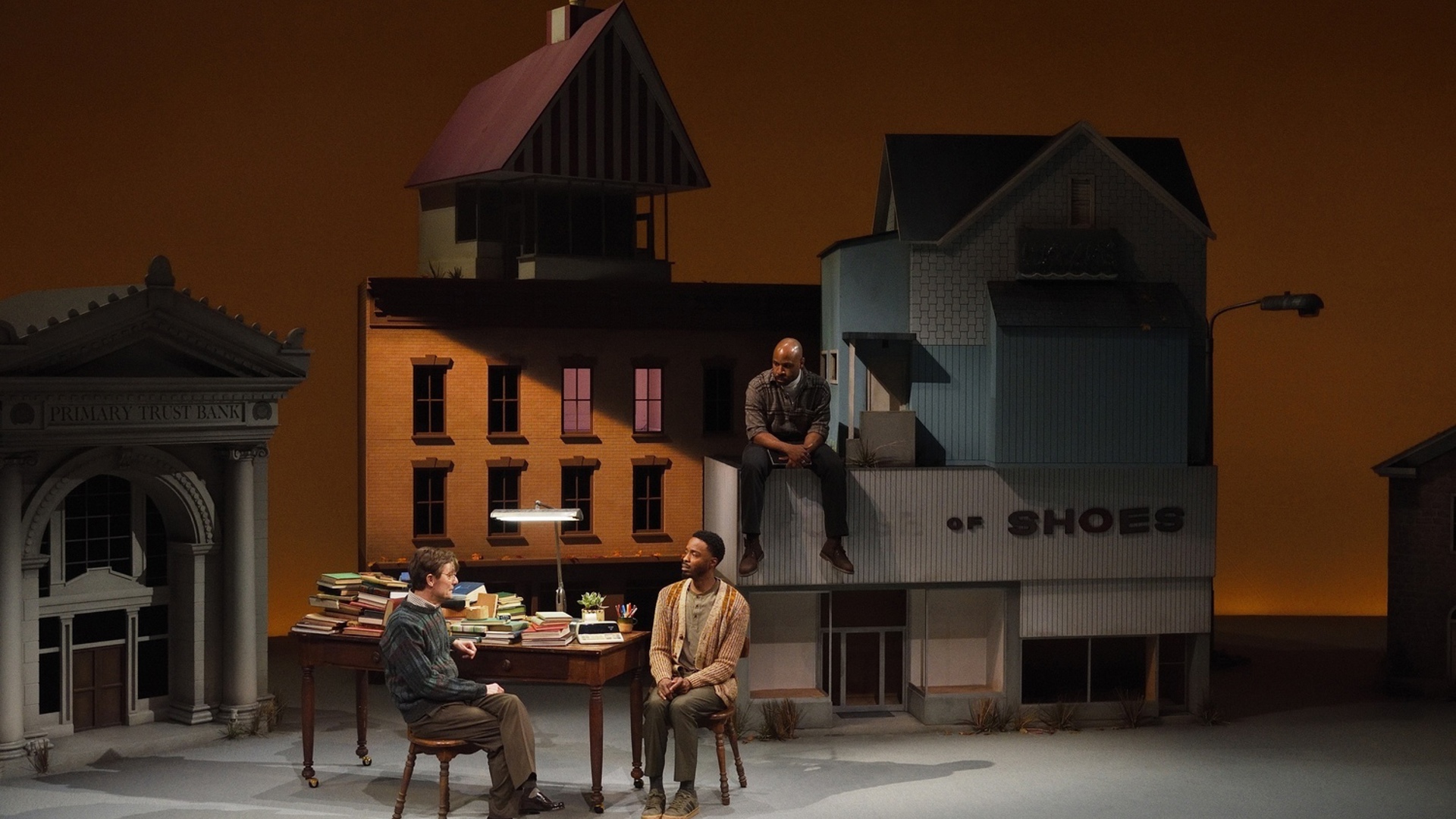 Two men sit a table covered with different books, while another man sits on the ledge of a building looking down at them in a town filled with large buildings.